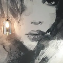 Ściana z fototapetą przedstawiającą abstrakcyjny portret kobiety w odcieniach szarości, oświetlona wiszącą lampą z przezroczystym kloszem i żarówką filamentową.