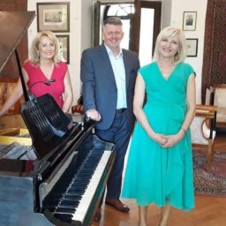 Arawak Krzysztof Boguń - Trzy osoby pozują obok czarnego fortepianu w pomieszczeniu z obrazami na ścianach i dywanem na podłodze.