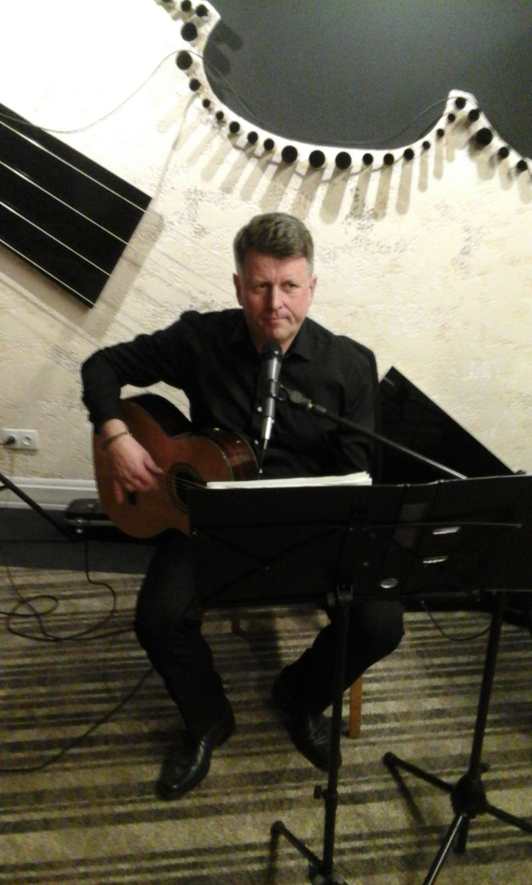 Mężczyzna w czarnym ubraniu gra na gitarze akustycznej przed mikrofonem i nutami, z dekoracją w kształcie gitary na ścianie w tle.