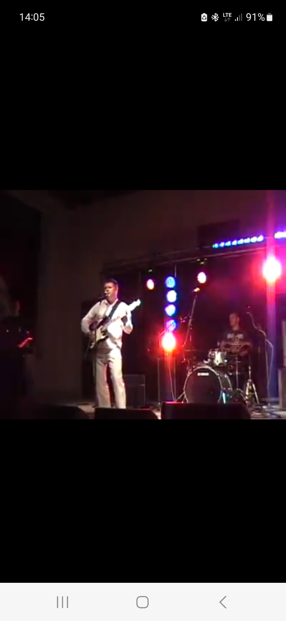 Zespół muzyczny grający na scenie podczas koncertu, wokalista z gitarą na pierwszym planie, perkusista w tle, oświetlenie sceniczne.