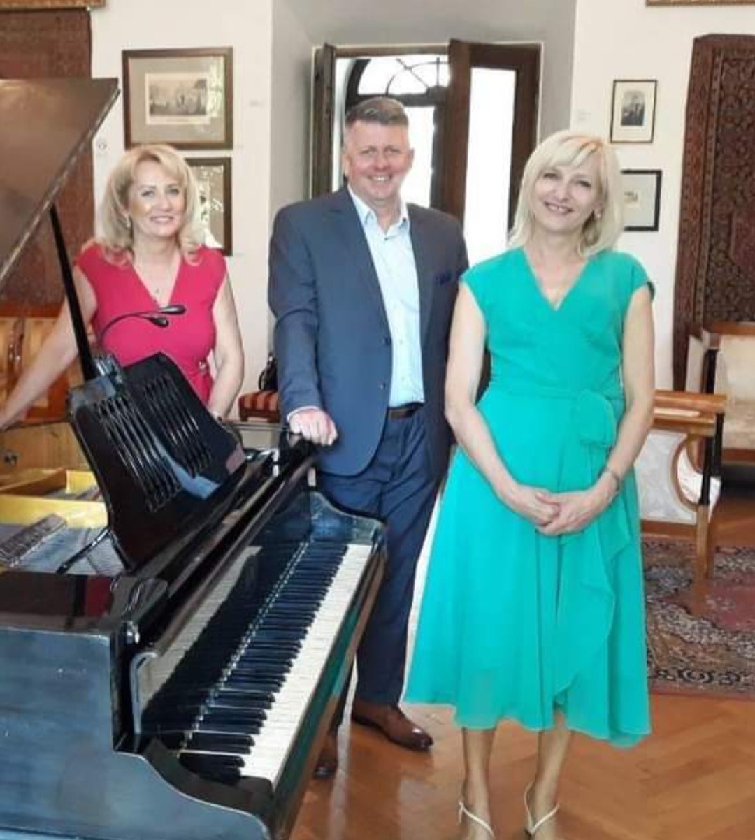 Trzy osoby pozują obok czarnego fortepianu w pomieszczeniu z obrazami na ścianach i dywanem na podłodze.