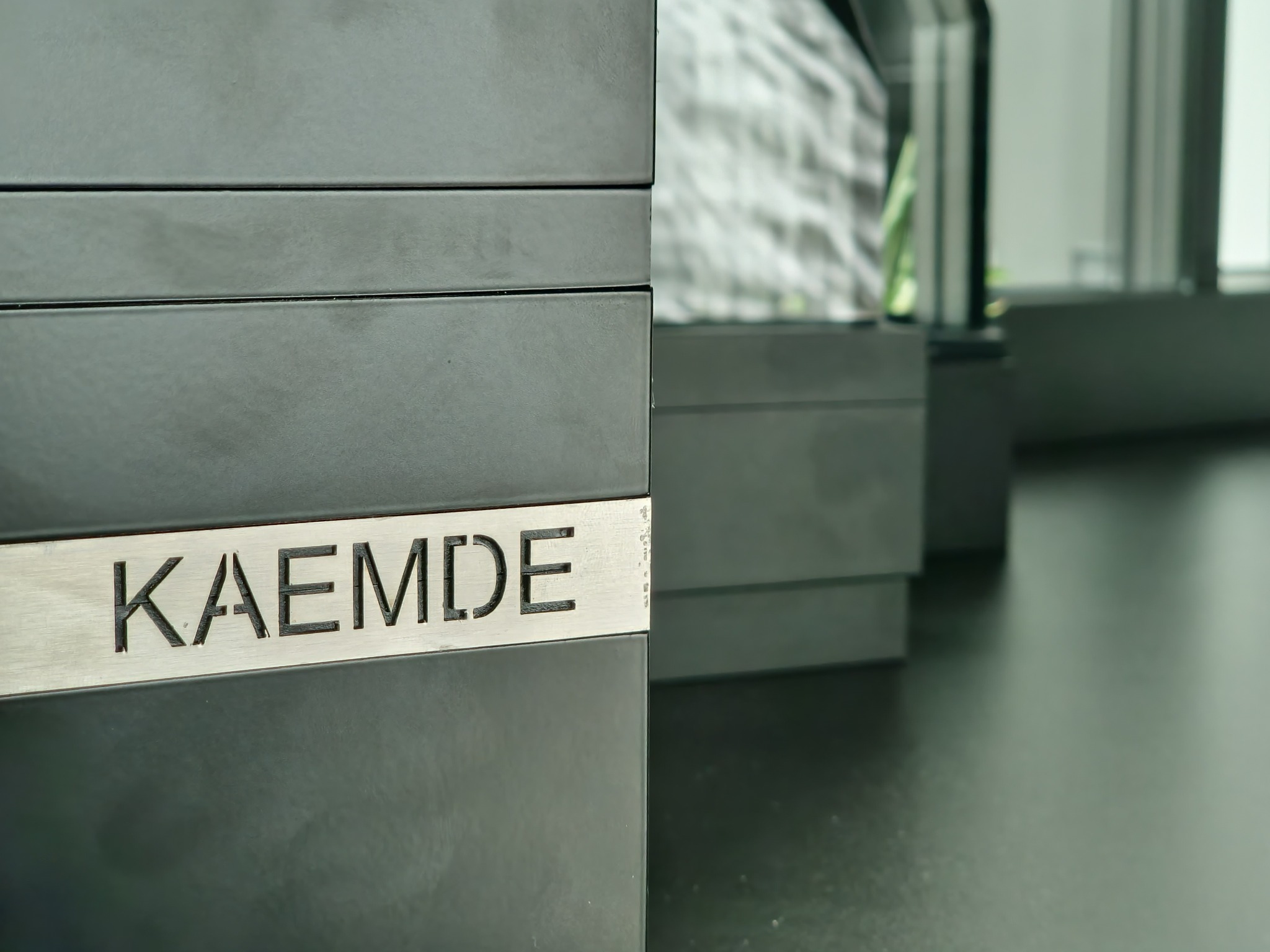 Ciemnoszare skrzydło drzwiowe z aluminiową tabliczką z wyciętym napisem 'KAEMDE', widoczne kolejne skrzydła drzwiowe i okno w tle.