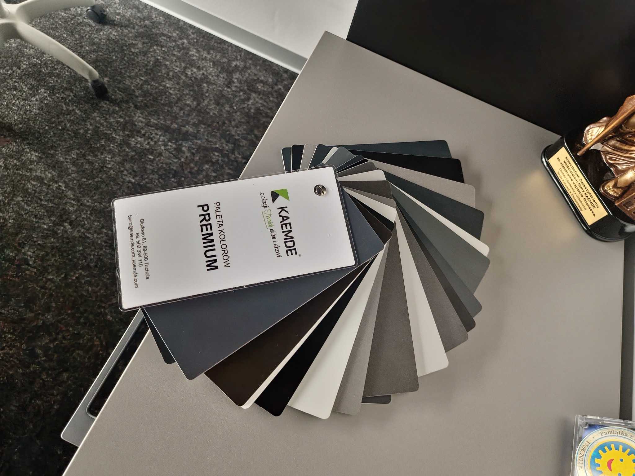 Paleta kolorów Premium firmy Kaemde, prezentująca odcienie szarości, leżąca na szarym blacie biurka obok statuetki z dedykacją.