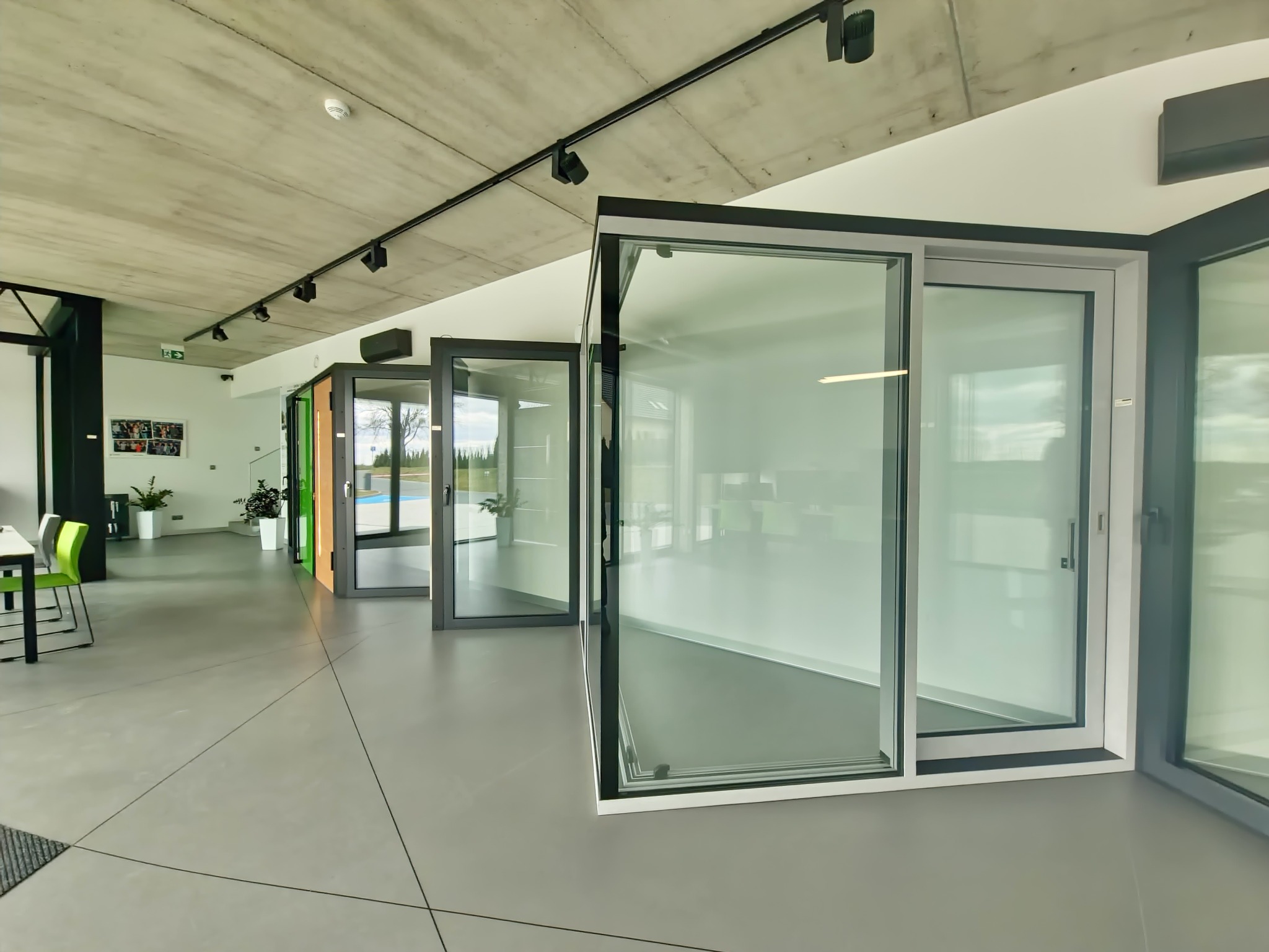 Wnętrze salonu z ekspozycją aluminiowych okien przesuwnych w szarych ramach, widok z perspektywy lekko z góry, betonowy sufit z oświetleniem szynowym.