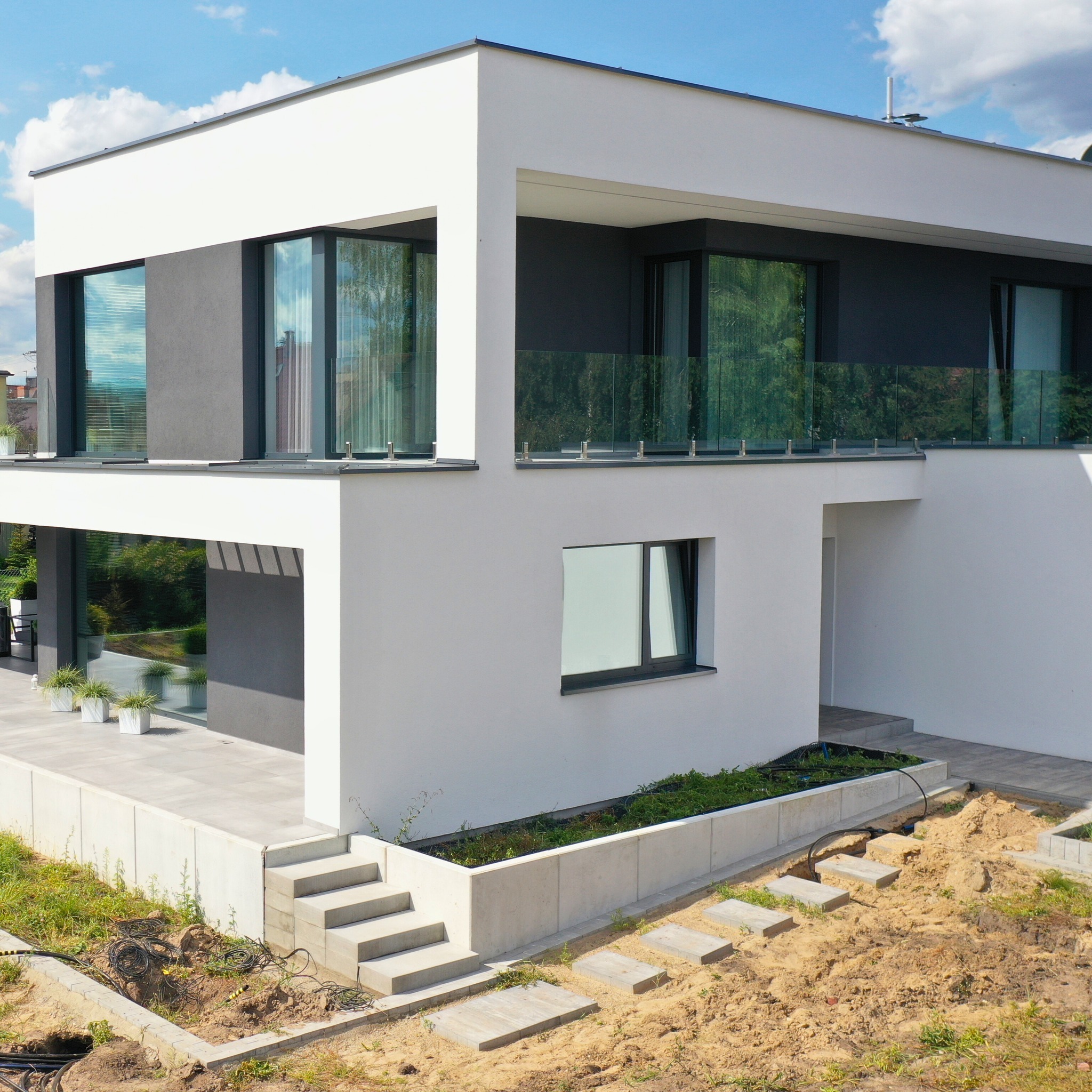 Nowoczesny dom z dużymi oknami aluminiowymi i szklaną balustradą na balkonie, otoczony surowym terenem z widocznymi kablami i betonowymi płytami chodnikowymi.