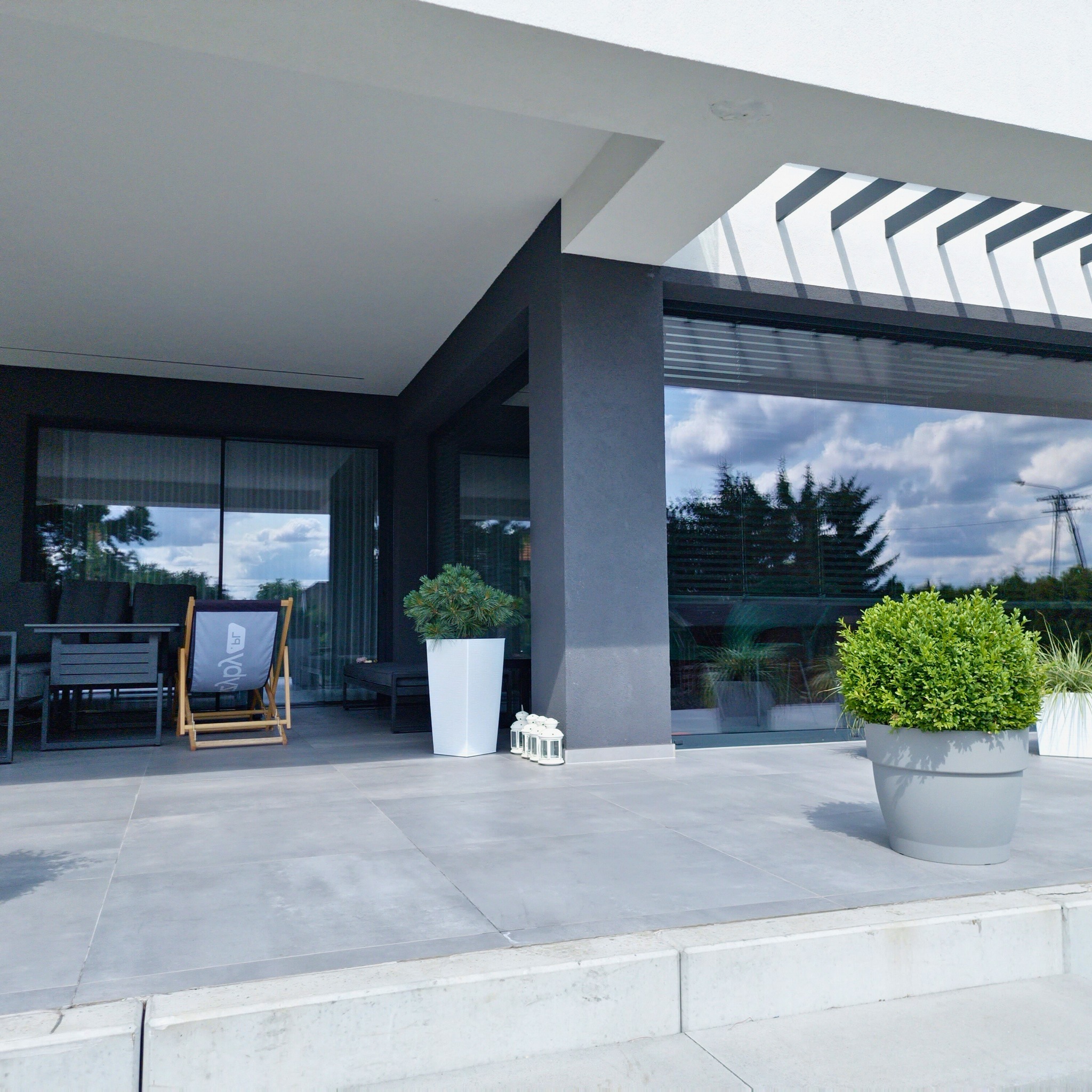 Nowoczesny taras z szarymi płytkami, drewnianym leżakiem z logo, roślinami w donicach i dużymi przesuwnymi oknami od podłogi do sufitu.