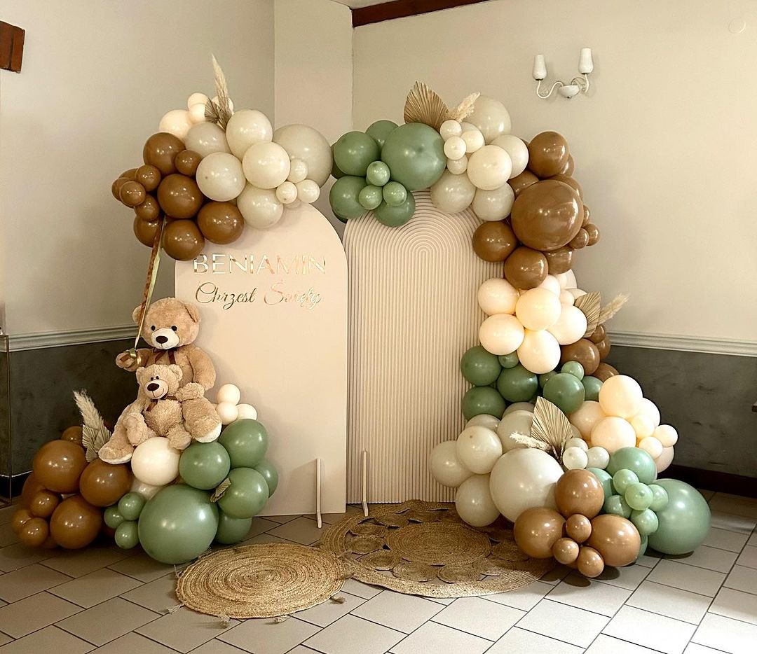 Dekoracja balonowa w odcieniach brązu, zieleni i bieli z pluszowymi misiami i napisem 'Benjamin Chrzest Święty' na tle beżowej ścianki z łukami.