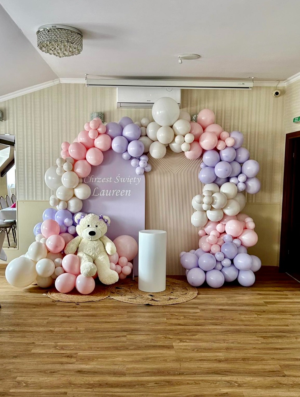 Dekoracja balonowa w odcieniach różu, fioletu i bieli z pluszowym misiem na tle ścianki z napisem 'Chrzest Święty Laureen' i plecionego dywanika.