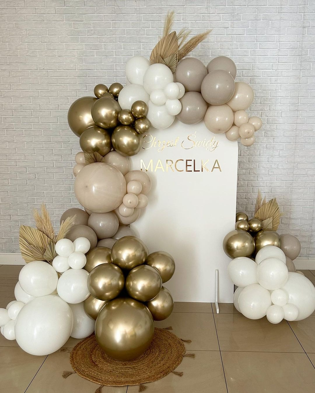 Dekoracja balonowa w odcieniach bieli, beżu i złota z napisem 'Chrzest Święty Marcelka' na białym tle, uzupełniona suszonymi trawami.