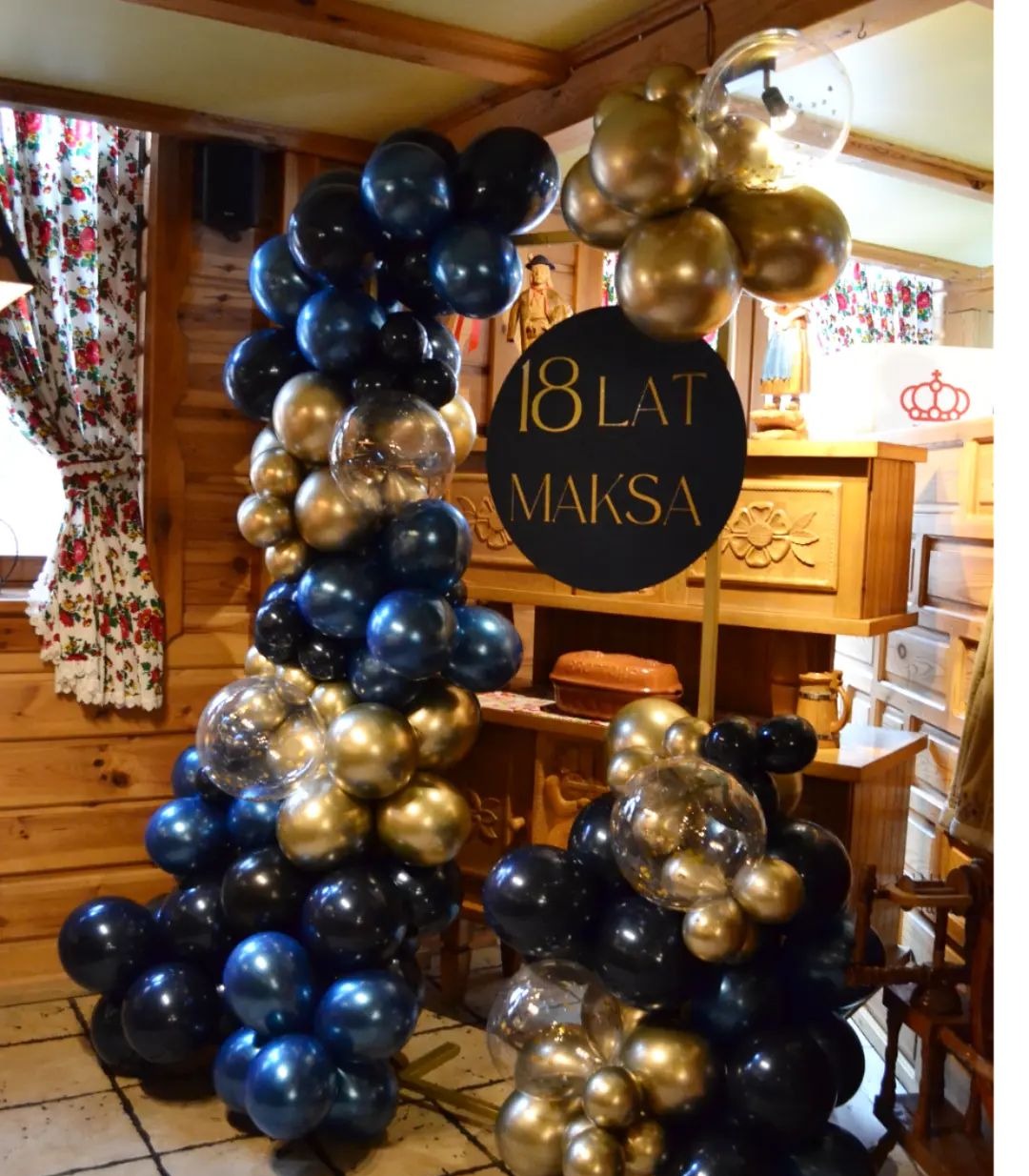 Dekoracja balonowa w kolorach granatu i złota z napisem '18 lat Maksa' na czarnym kole, ustawiona w drewnianym wnętrzu.