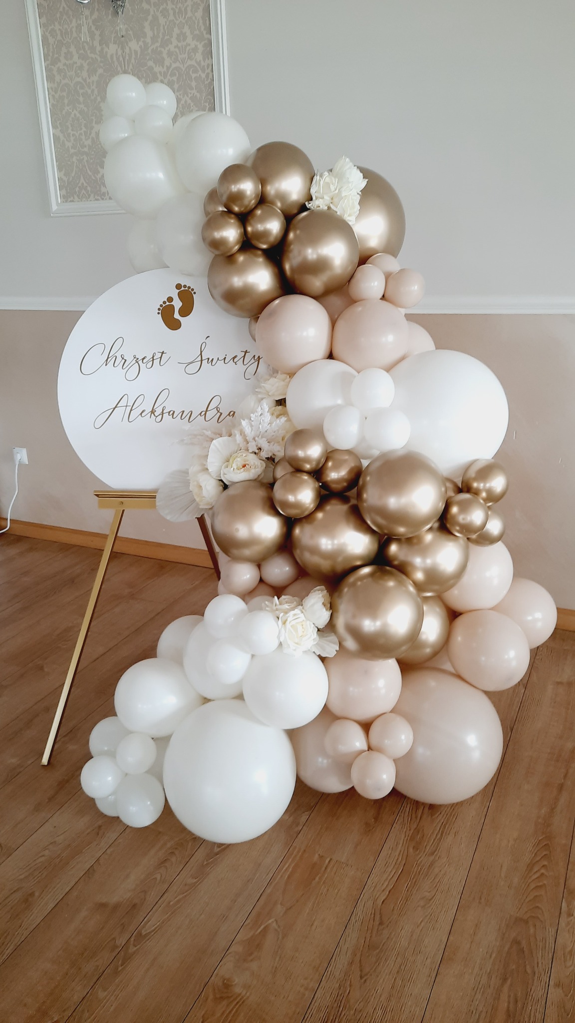 Dekoracja balonowa w odcieniach bieli, beżu i złota, z białymi kwiatami, umieszczona obok okrągłej tablicy z napisem 'Chrzest Święty Aleksandra' i złotymi odciskami stóp, na złotym stojaku.