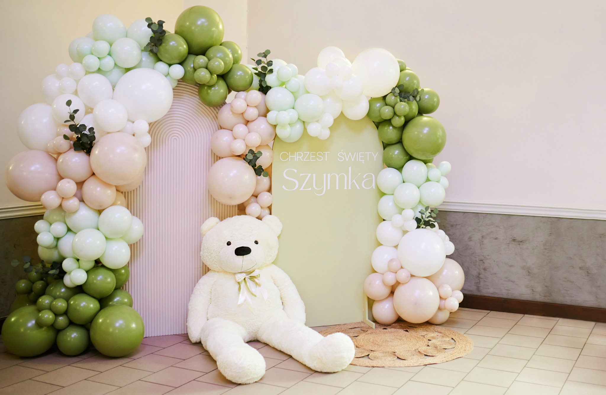 Dekoracja balonowa w odcieniach zieleni, bieli i różu z napisem 'Chrzest Święty Szymka', uzupełniona pluszowym misiem i okrągłym jutowym dywanikiem.