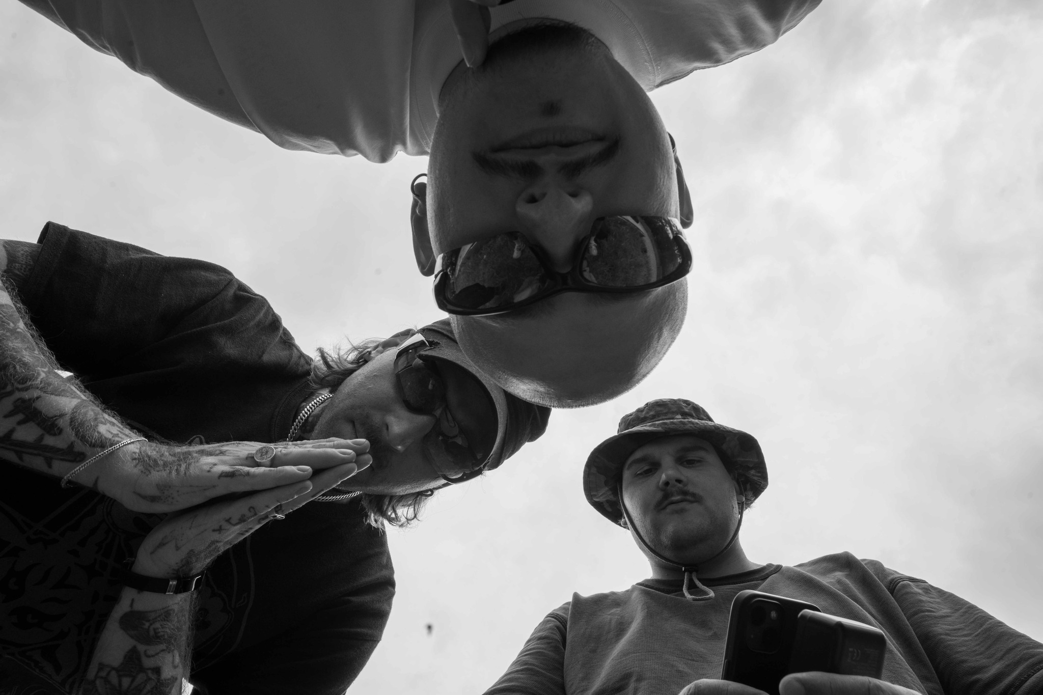 Czarno-białe ujęcie z dołu przedstawiające trzech mężczyzn, jeden z nich odwrócony do góry nogami w okularach przeciwsłonecznych, drugi z tatuażami na rękach, a trzeci w kapeluszu trzymający telefon.