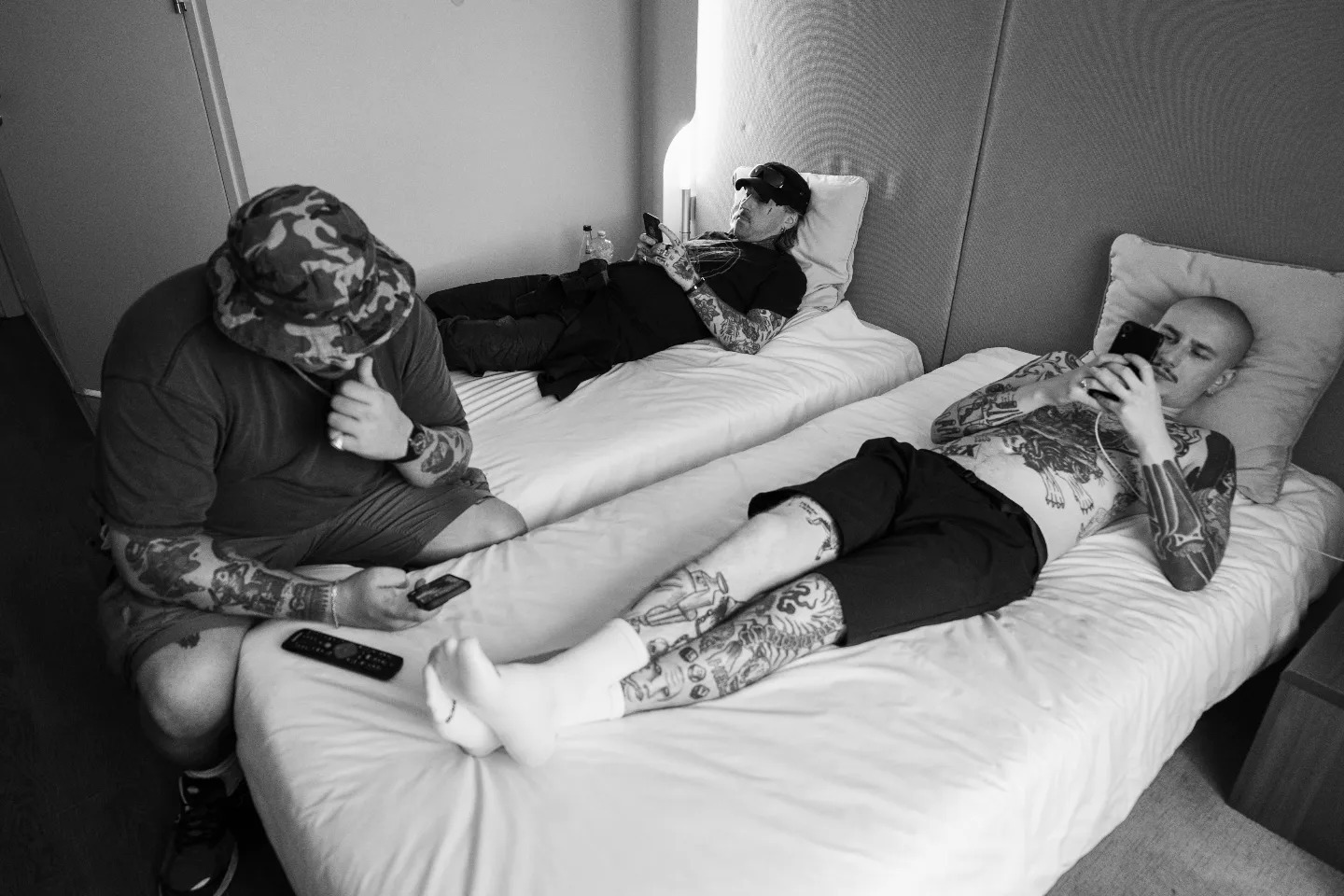 Czarno-białe zdjęcie trzech wytatuowanych mężczyzn odpoczywających na łóżkach w pokoju hotelowym, każdy z nich używa telefonu komórkowego.