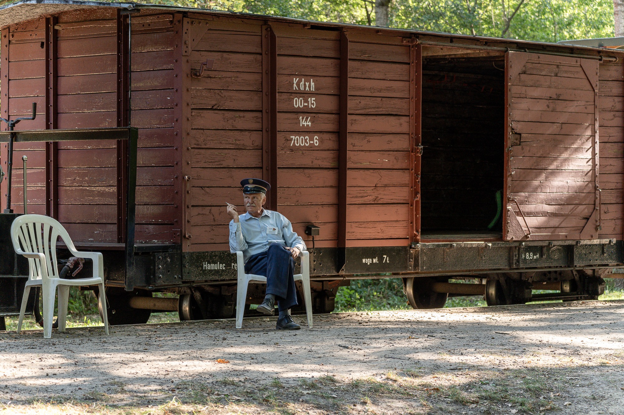 Starszy mężczyzna w mundurze siedzi na plastikowym krześle przed otwartym, drewnianym wagonem kolejowym, paląc papierosa.