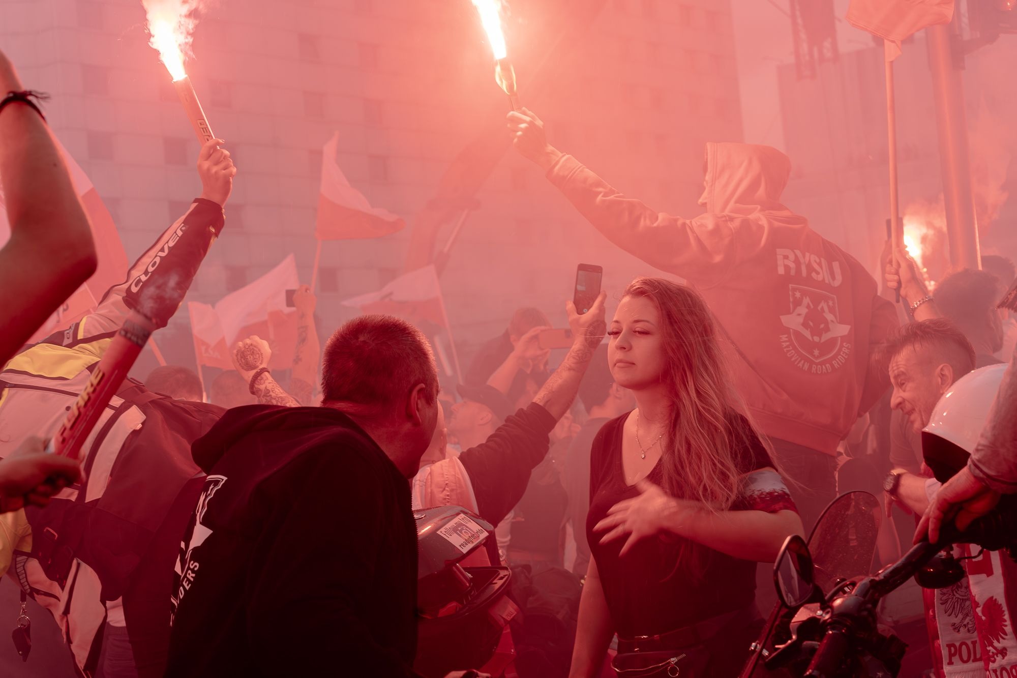 Tłum ludzi z polskimi flagami i racami podczas wydarzenia plenerowego, kobieta w centrum kadru z zamyślonym wyrazem twarzy, czerwona poświata od rac.