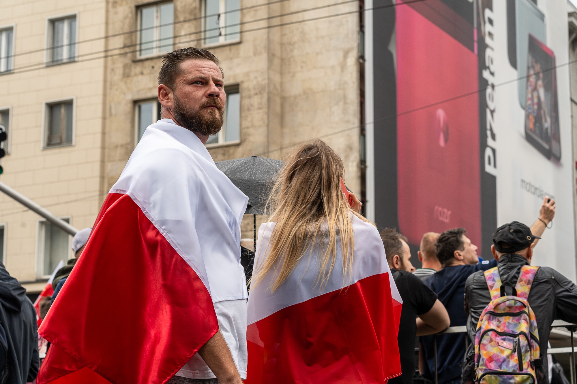 Mężczyzna z brodą i blondwłosa kobieta owinięci flagami Polski, patrzący w górę na tle budynku i reklamy telefonu.