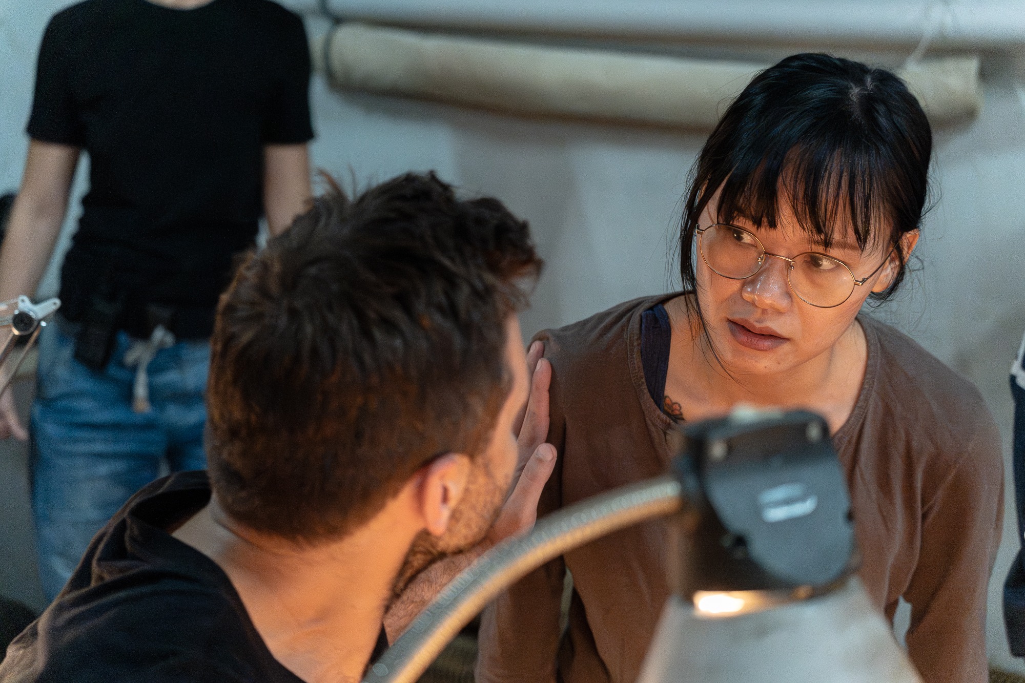 Kobieta w okularach z zaciekawieniem spogląda na mężczyznę podczas nagrywania filmu, widoczny sprzęt oświetleniowy i fragment kadru zza pleców operatora