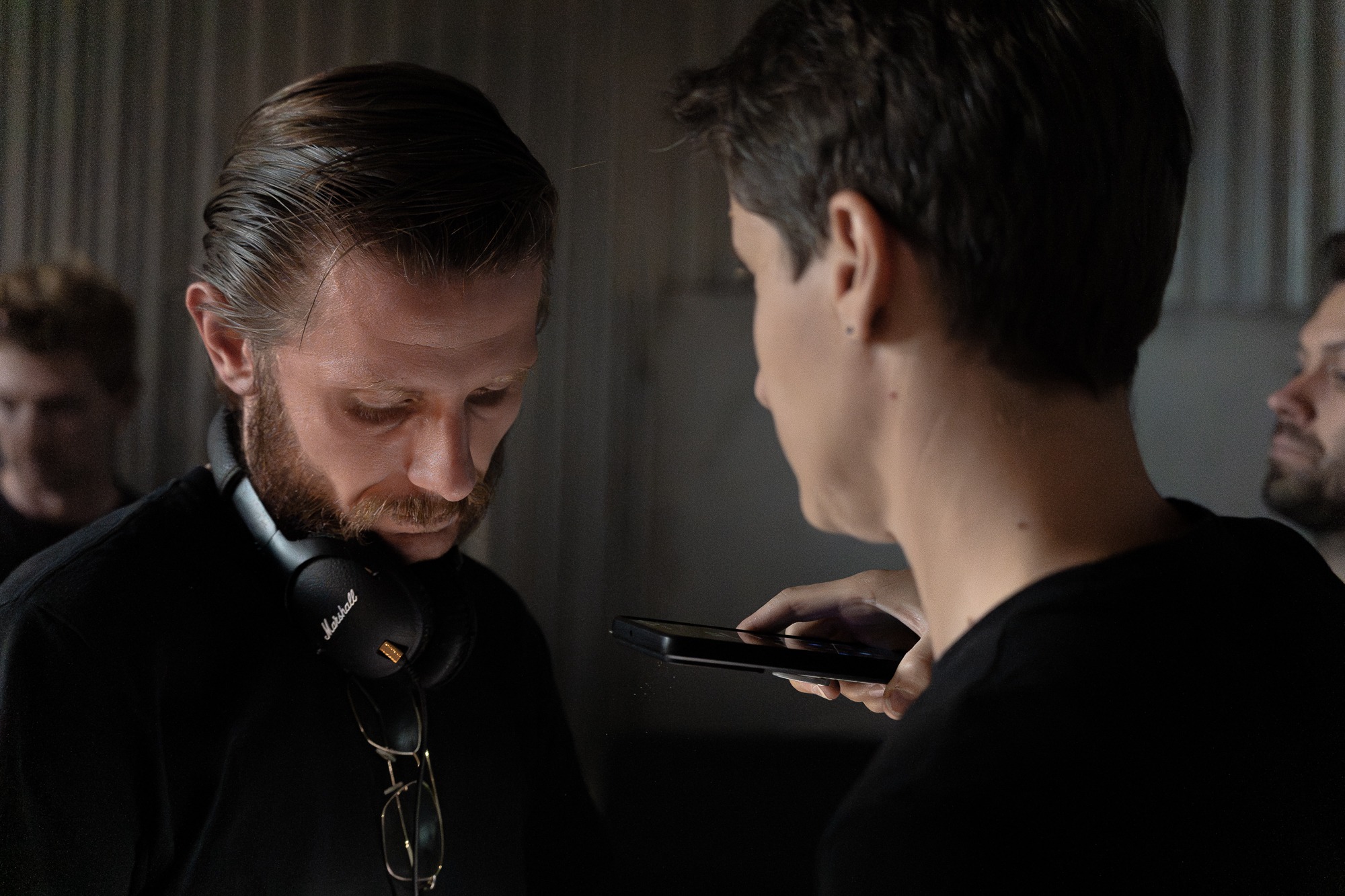 Kamerzysta z brodą i słuchawkami Marshall na szyi, omawia ujęcia z asystentem trzymającym smartfon, w tle widoczni inni członkowie ekipy.