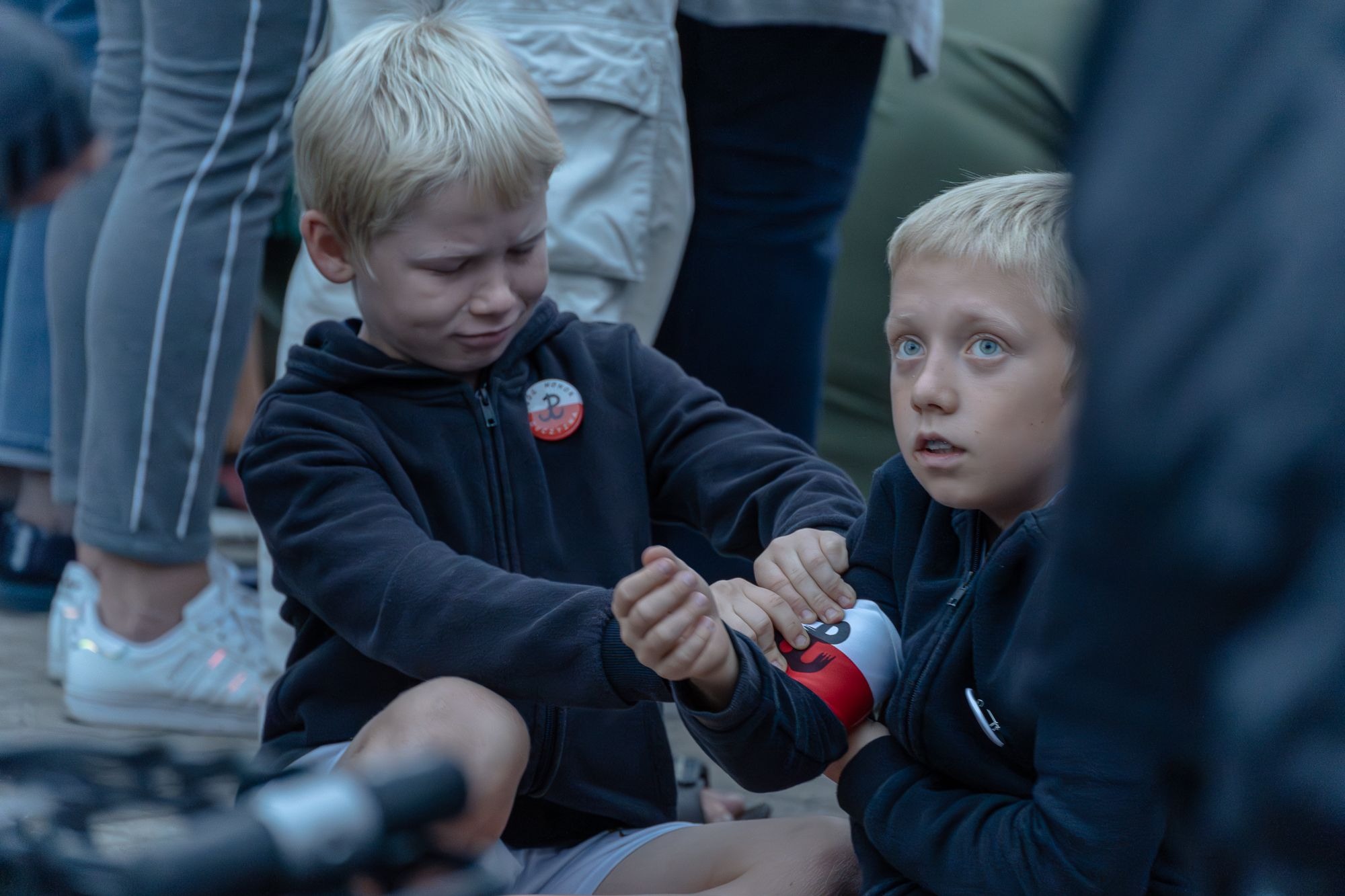 Dwóch blond chłopców w bluzach z godłem Polski siedzi na ziemi, jeden z grymasem bólu, drugi z wyrazem zaniepokojenia, trzymając bandaż na ramieniu kolegi.