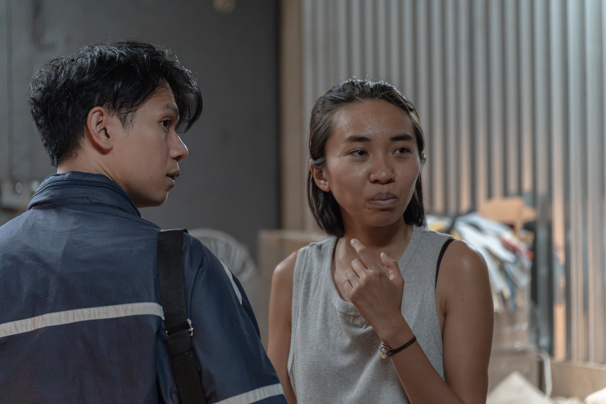 Dwoje młodych ludzi w pomieszczeniu o surowym wystroju, kobieta w szarej koszulce bez rękawów z bransoletką na nadgarstku gestykuluje, mężczyzna w granatowej kurtce z torbą na ramieniu patrzy w jej...