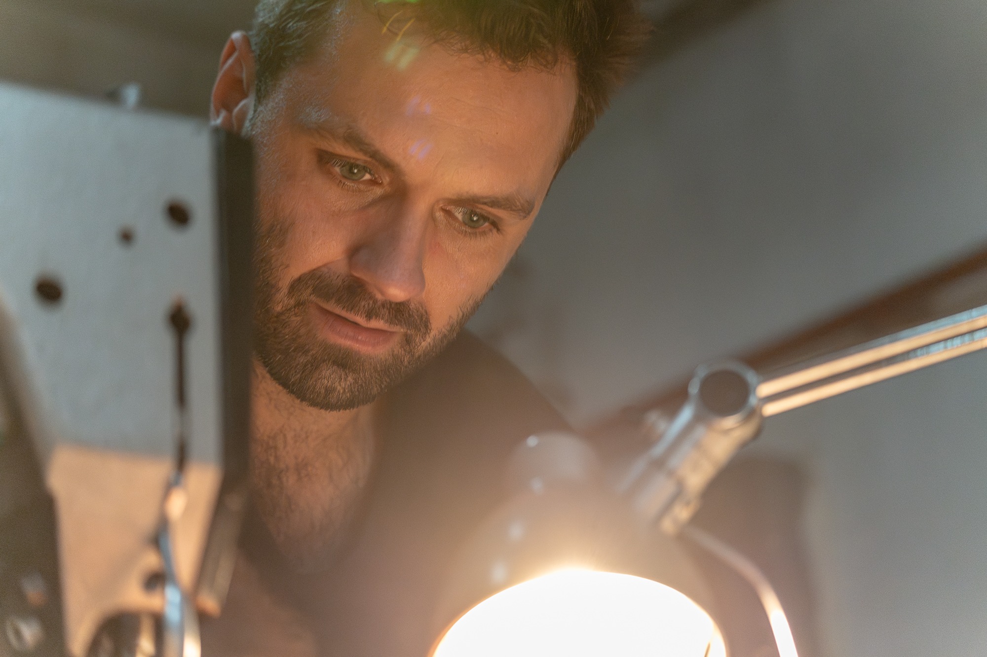 Portret mężczyzny z brodą, skupionego na pracy przy maszynie do szycia w oświetleniu lampy warsztatowej.