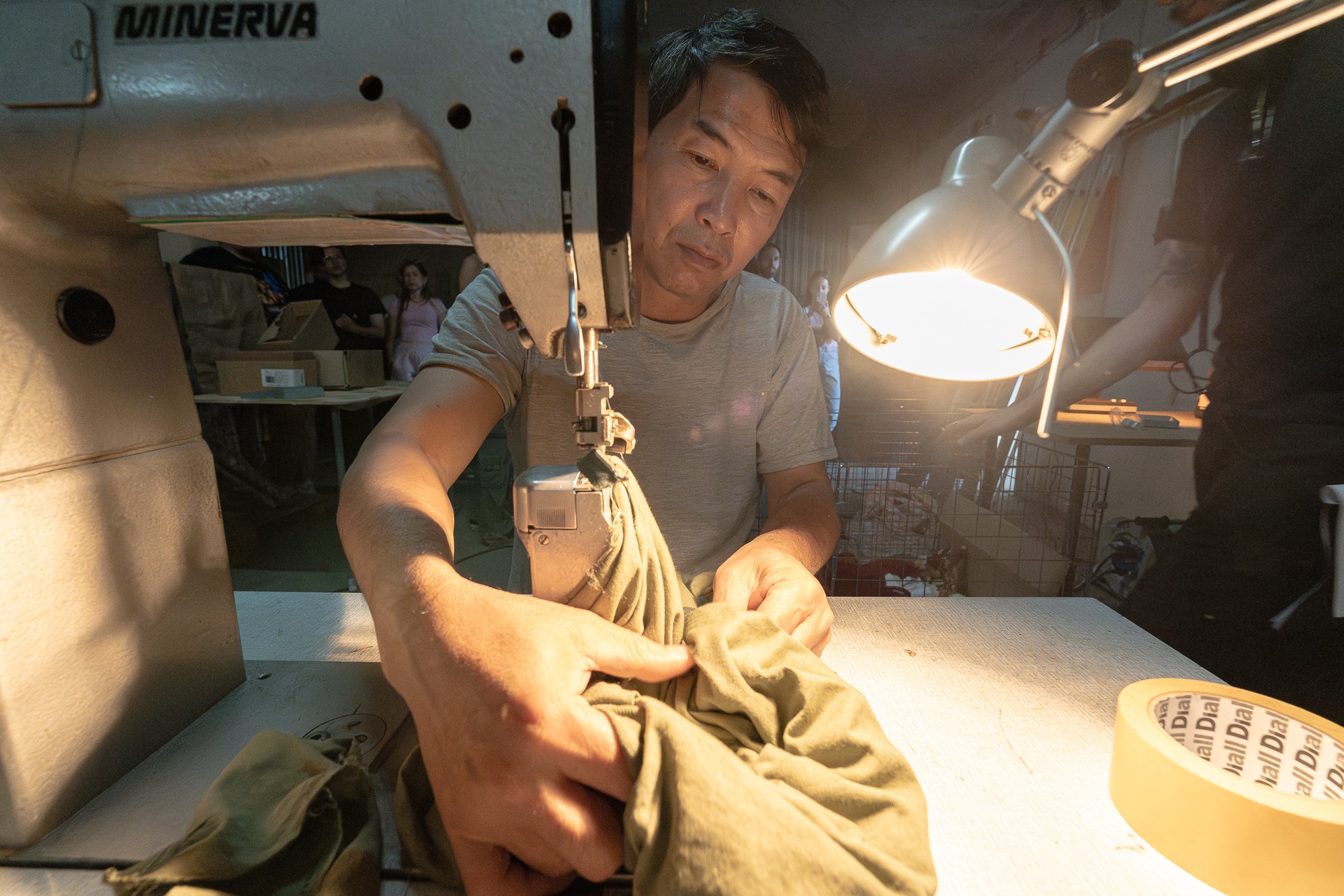 Szczegółowe ujęcie krawca przy pracy na maszynie do szycia Minerva, skupionego na zszywaniu zielonej tkaniny pod jasnym światłem lampki biurkowej, z widoczną taśmą klejącą na stole i postaciami w tle.