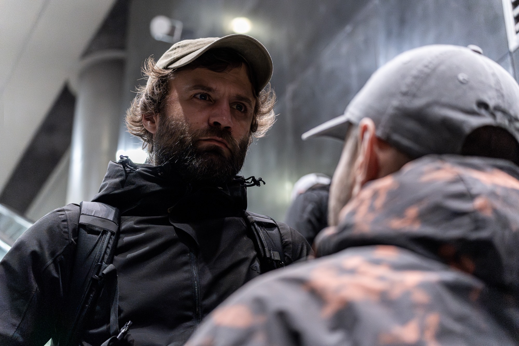 Mężczyzna z brodą i w czapce patrzy w górę na osobę z czapką, widoczne schody ruchome w tle.