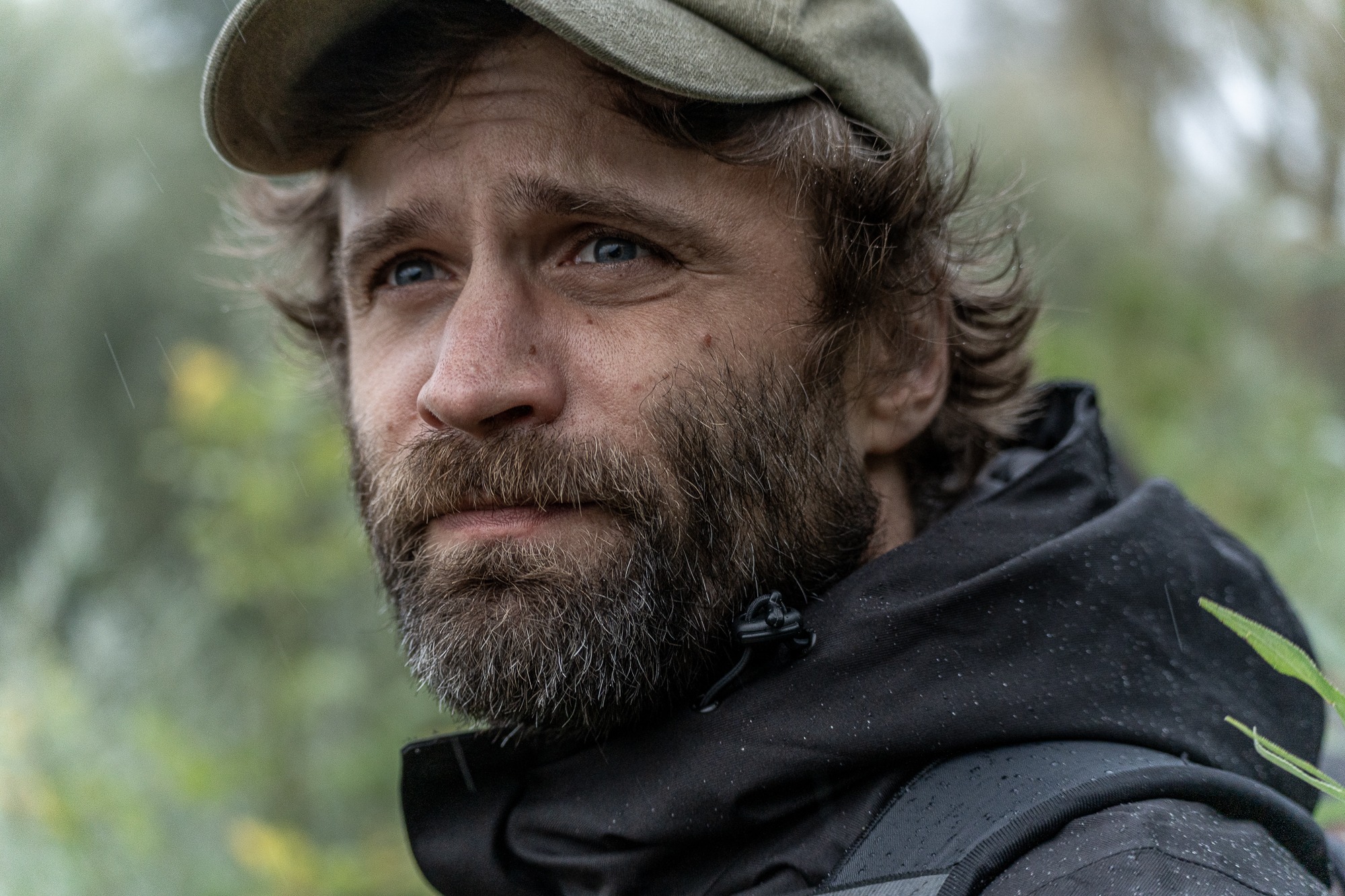 Portret brodatego mężczyzny w czapce, z kroplami deszczu na ubraniu, zamyślony wzrok skierowany w górę, tło zamazane, zielona roślinność.