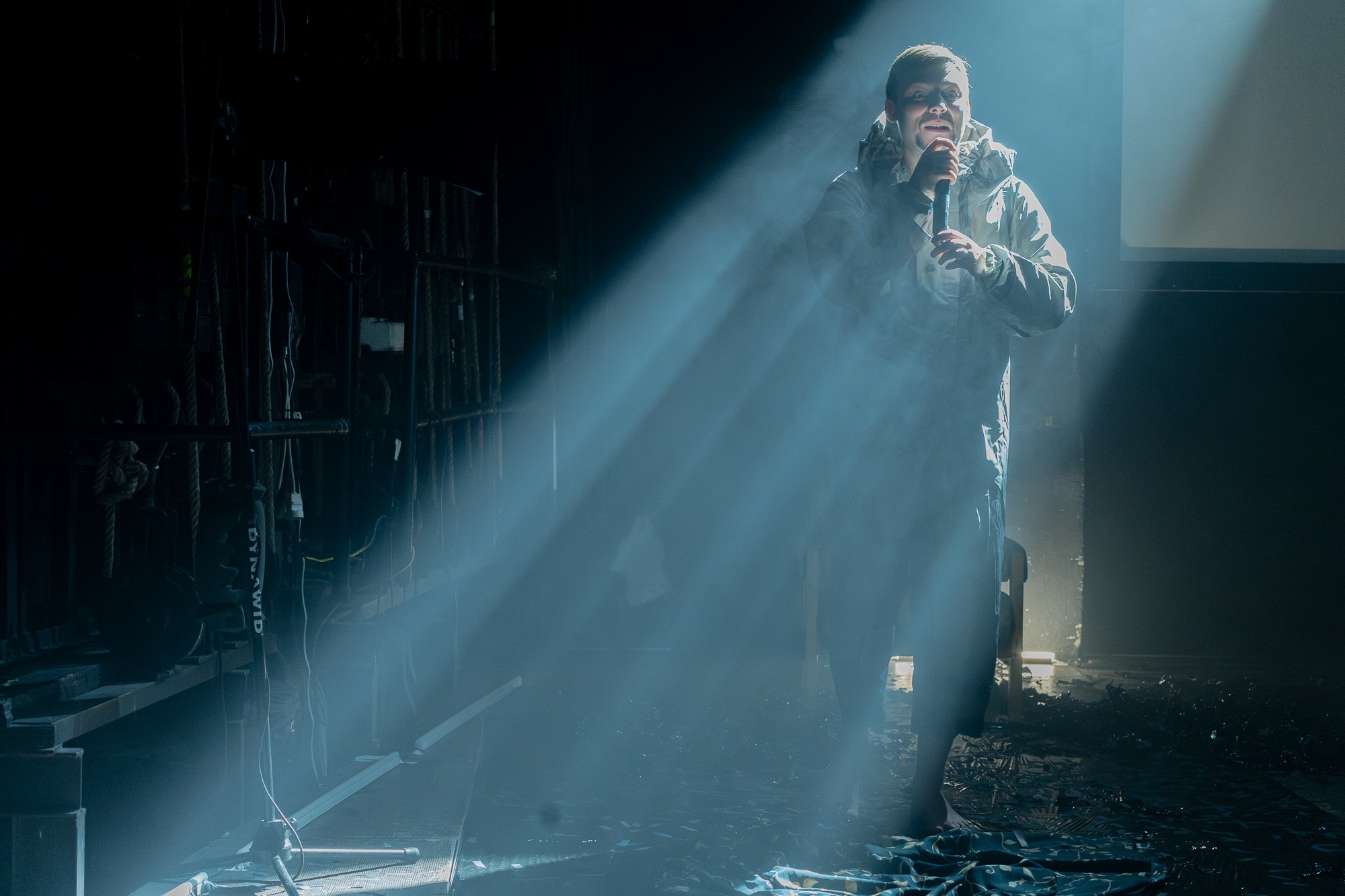 Mężczyzna w kurtce z kapturem stoi na scenie w snopie światła, trzymając mikrofon. Scena jest zagracona kablami i sprzętem.