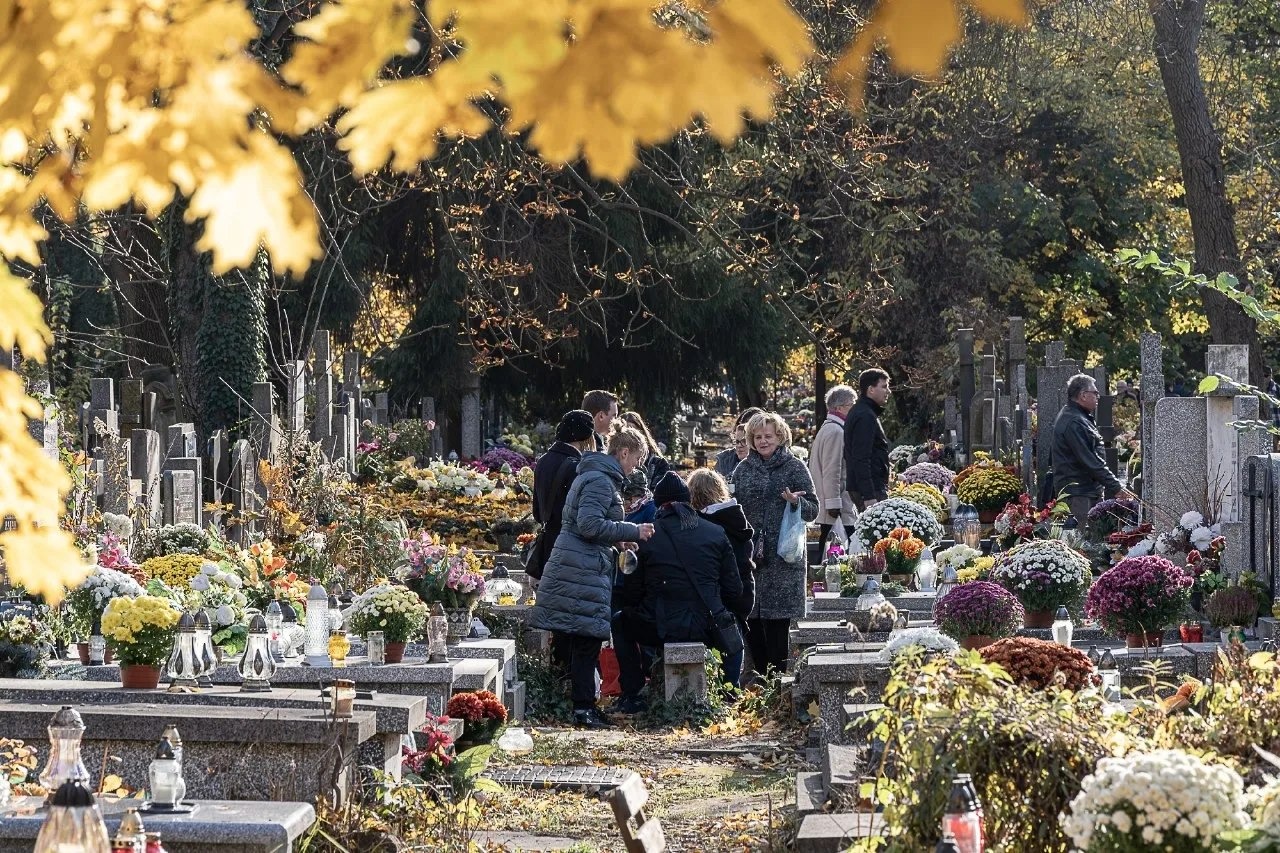 Jesienny krajobraz cmentarza z nagrobkami ozdobionymi kwiatami i zniczami, gdzie w tle widać grupę ludzi.