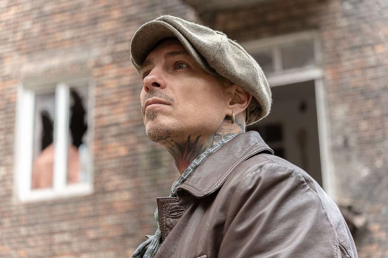 Mężczyzna w kaszkiecie i skórzanej kurtce, z tatuażami na szyi, spogląda w górę na tle ceglanego budynku z wybitymi oknami.