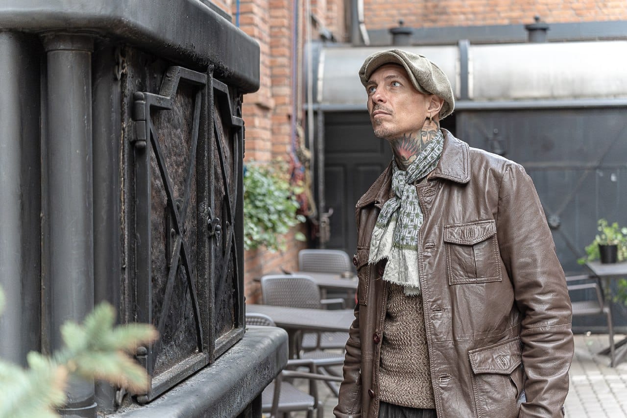 Mężczyzna w skórzanej kurtce, czapce i szaliku z tatuażami na szyi, stojący na zewnątrz obok metalowej konstrukcji, spoglądający w górę.