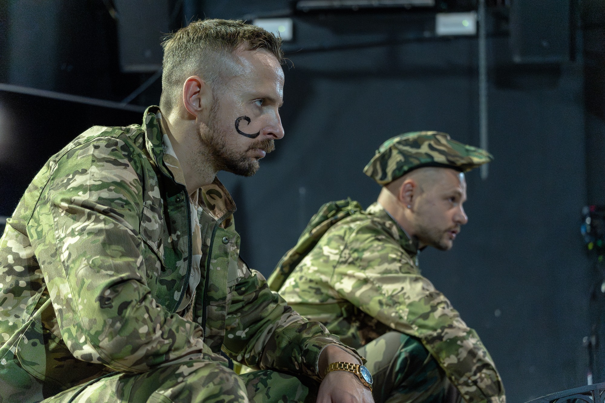 Dwóch mężczyzn w strojach moro, jeden z namalowanym wąsem na twarzy, siedzi na scenie w ciemnym otoczeniu.