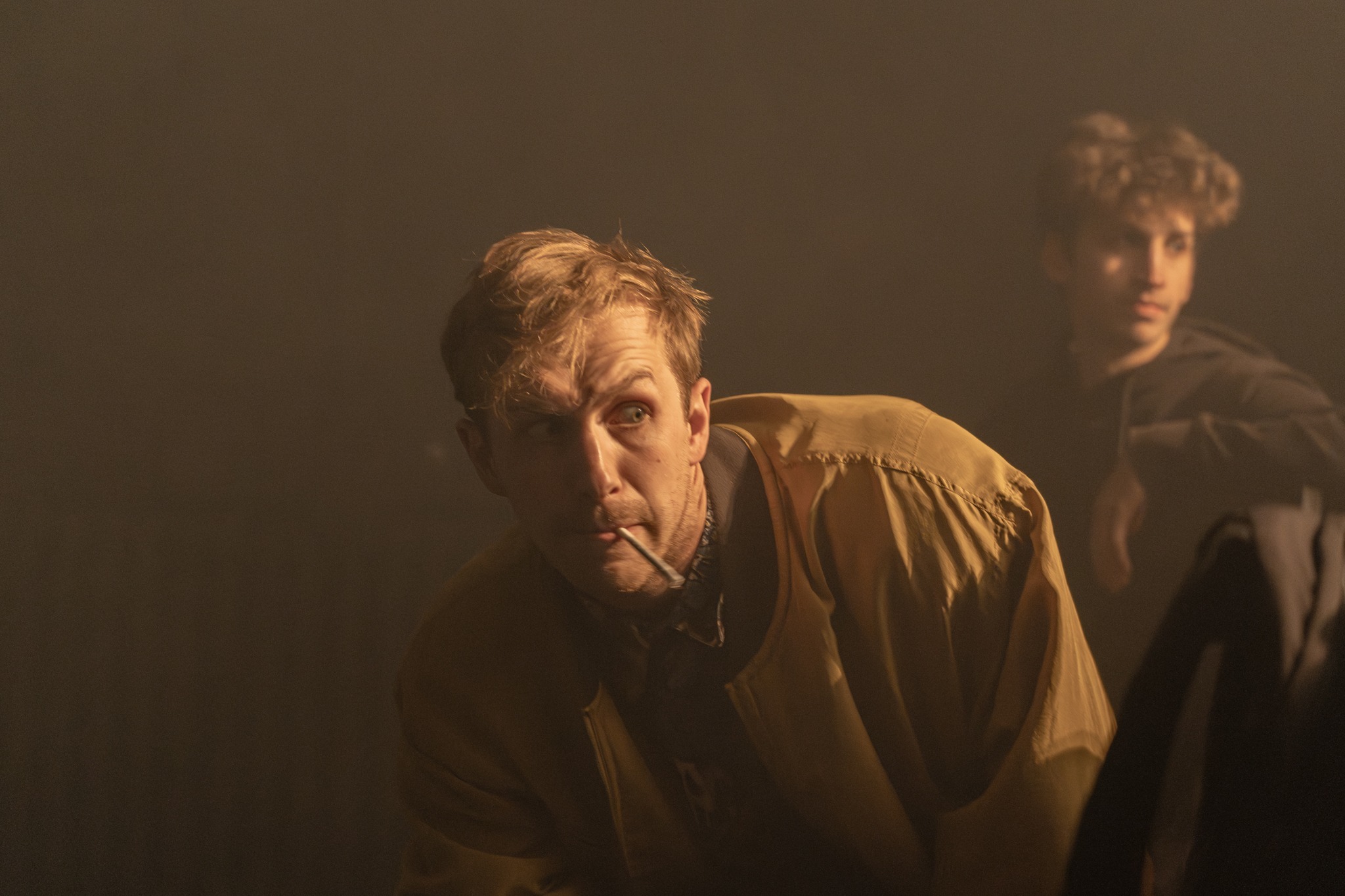 Mężczyzna z papierosem w ustach, ubrany w kurtkę, spogląda w górę w dymnym otoczeniu, w tle widoczna rozmazana postać młodego mężczyzny.