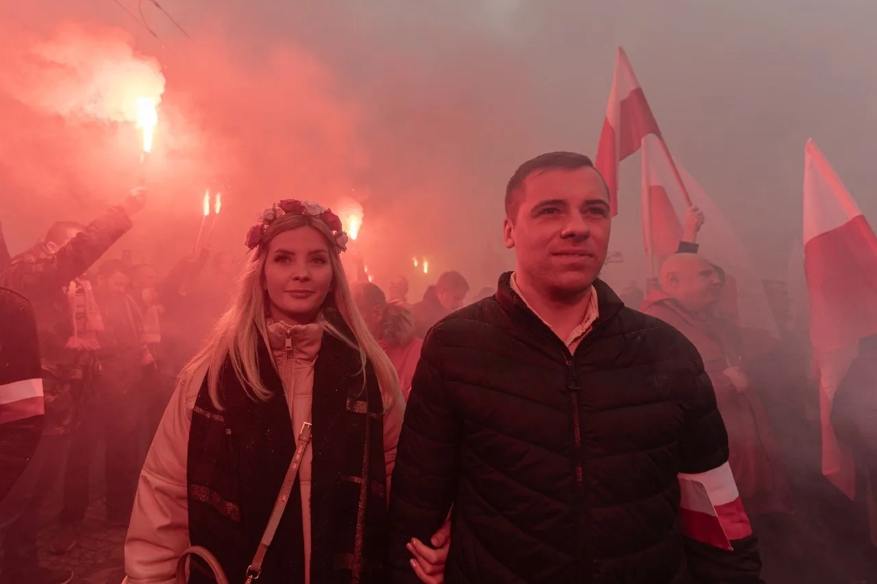 Para trzymająca się za ręce w tłumie podczas marszu z racami i flagami Polski. Kobieta ma wianek z kwiatów na głowie, a mężczyzna opaskę z flagą Polski na ramieniu.