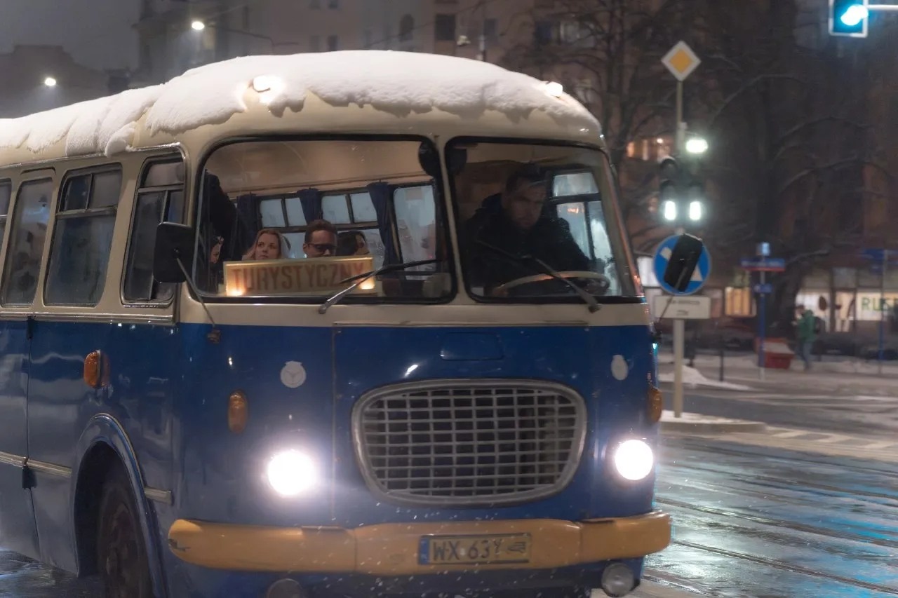 Niebiesko-biały autobus turystyczny z napisem 'Turystyczny' za szybą, pokryty śniegiem, przejeżdża przez zasypaną śniegiem ulicę Warszawy w zimowy wieczór.