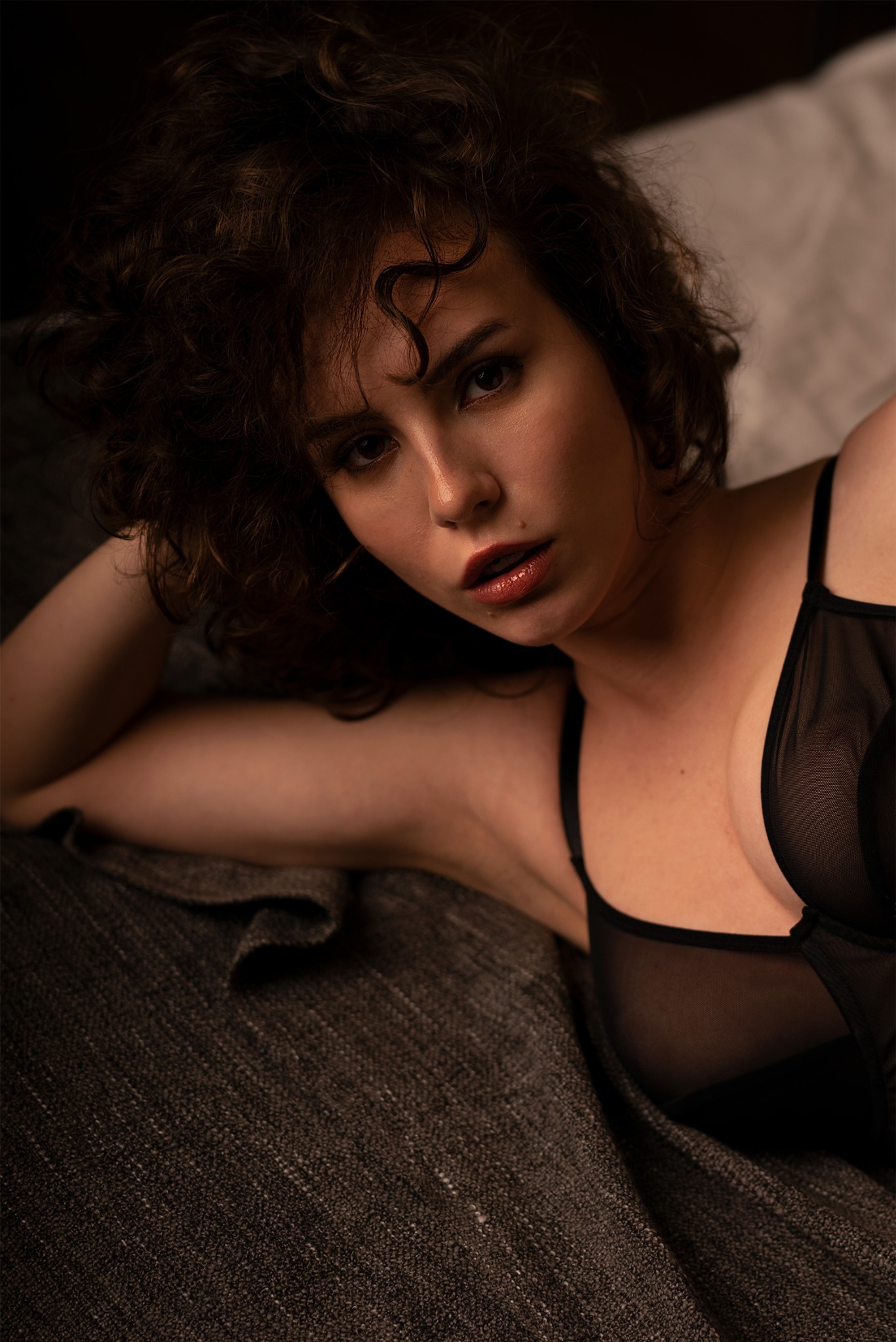Portret kobiety z kręconymi włosami w czarnej bieliźnie na tle ciemnoszarej pościeli, wykonany w Chojnowie w klimacie 'sesji sensual'.