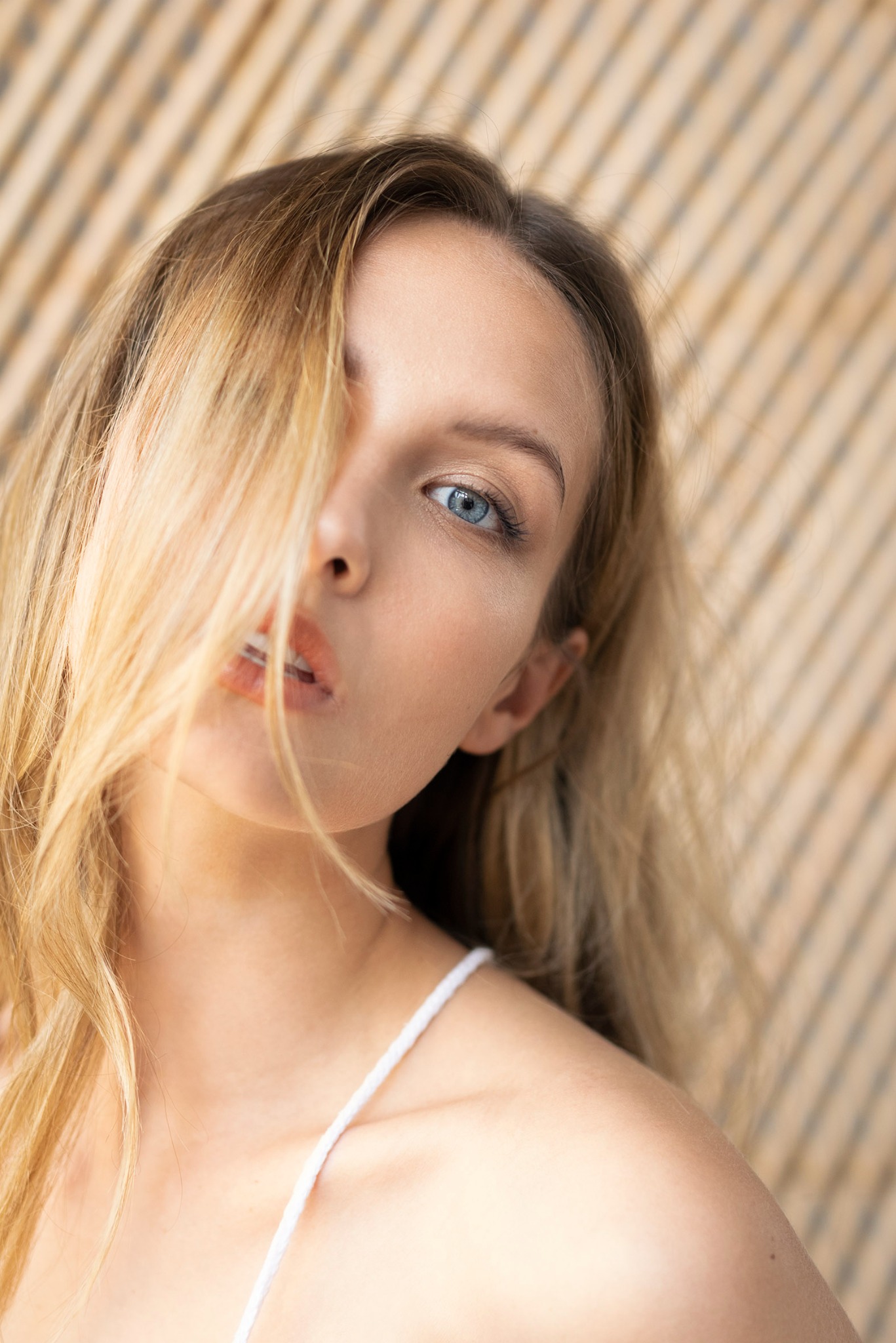 Portret kobiety z blond włosami przesłaniającymi część twarzy, błękitne oko w centrum uwagi, delikatny makijaż, drewniane tło w tle.