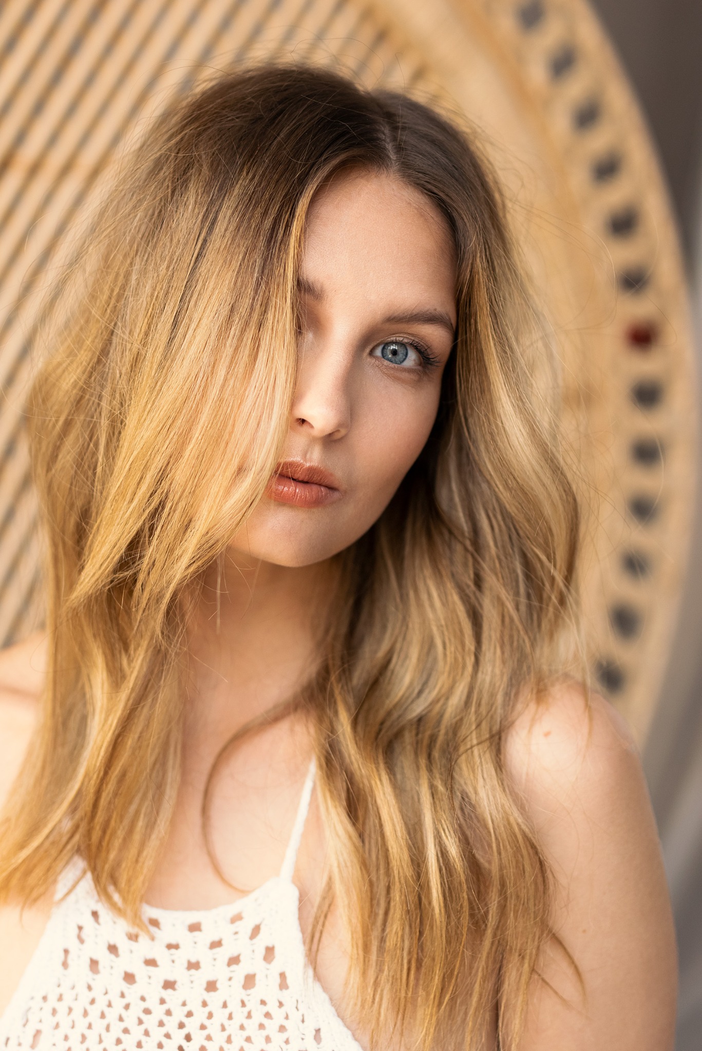 Portret kobiety z blond włosami zakrywającymi jedno oko, jasna karnacja, niebieskie oczy, w białej, ażurowej bluzce na tle dekoracyjnej ściany z drewna.