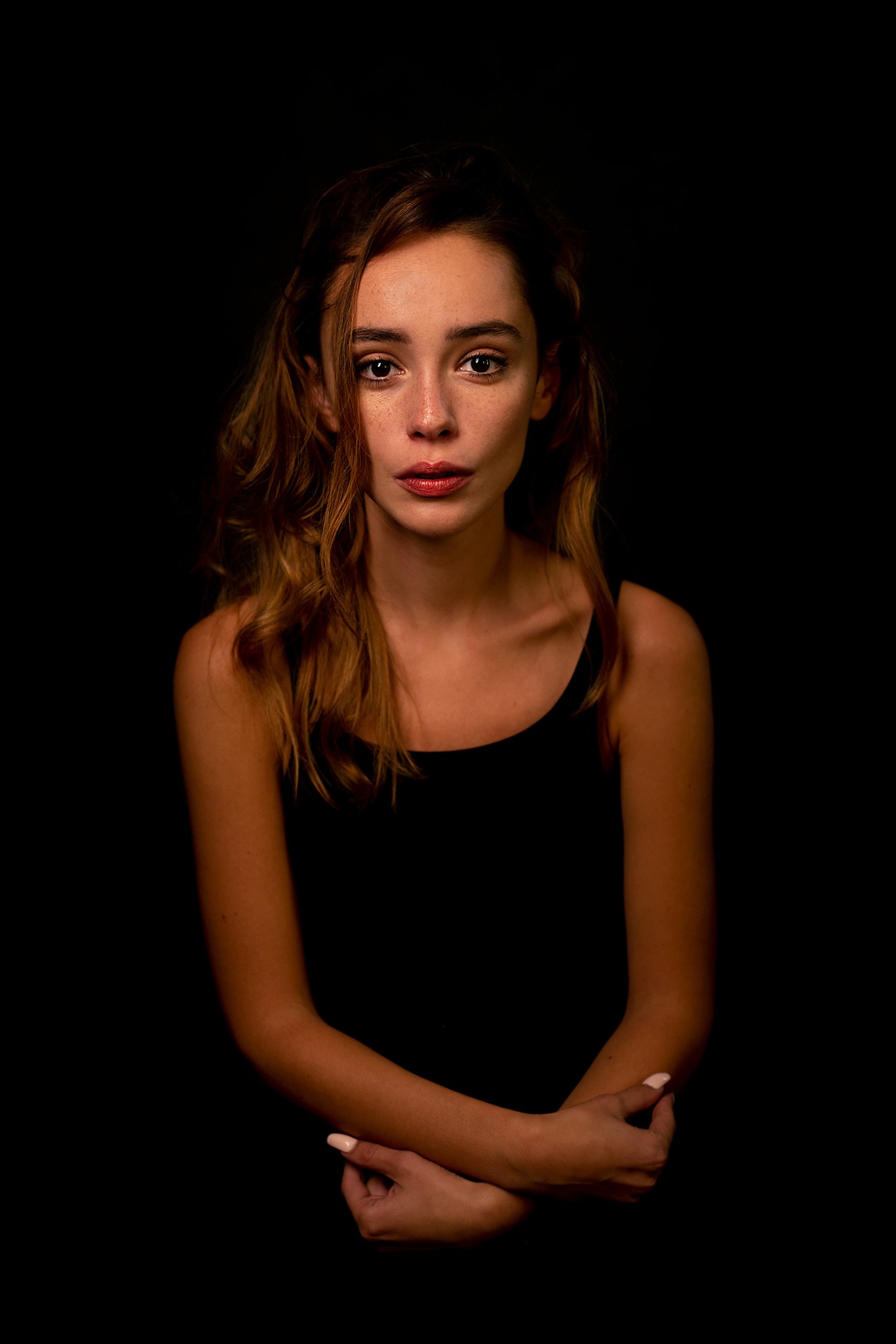 Portret kobiety z rudymi włosami i piegami, na ciemnym tle, z rękami skrzyżowanymi na wysokości klatki piersiowej.