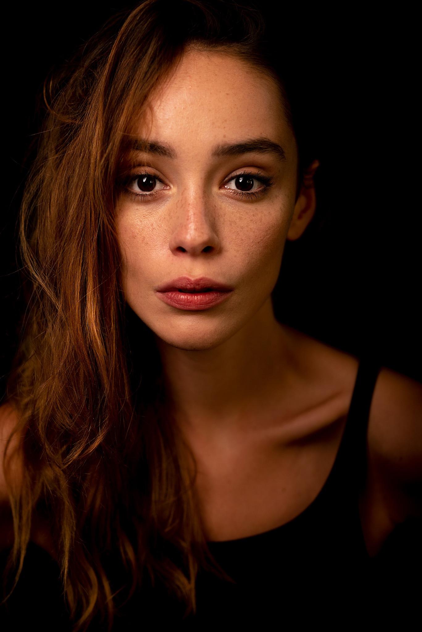 Portret młodej kobiety z piegami, z długimi, falowanymi, brązowymi włosami, spoglądającej w obiektyw na ciemnym tle.