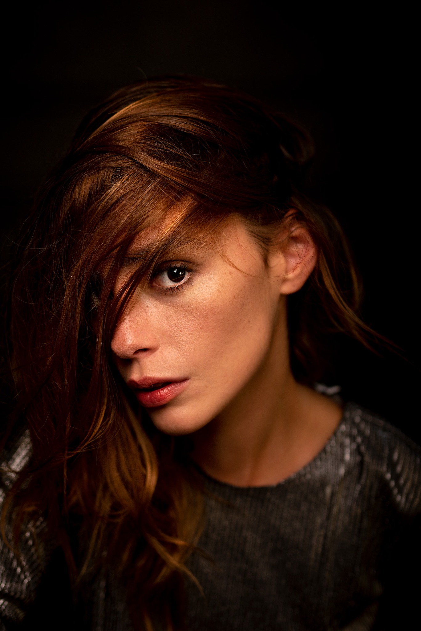 Portret kobiety z brązowymi włosami zasłaniającymi częściowo twarz, ciemne tło, oświetlenie boczne podkreślające rysy twarzy i piegi.
