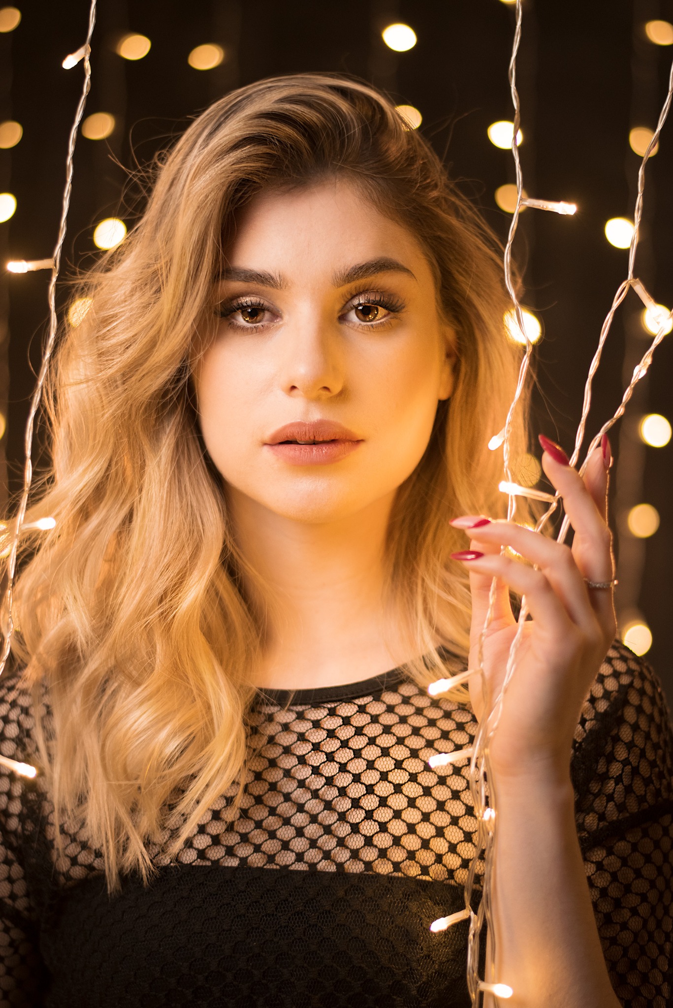 Portret kobiety z blond włosami i czerwonym manicure, trzymającej w dłoni światełka choinkowe na tle bokeh.