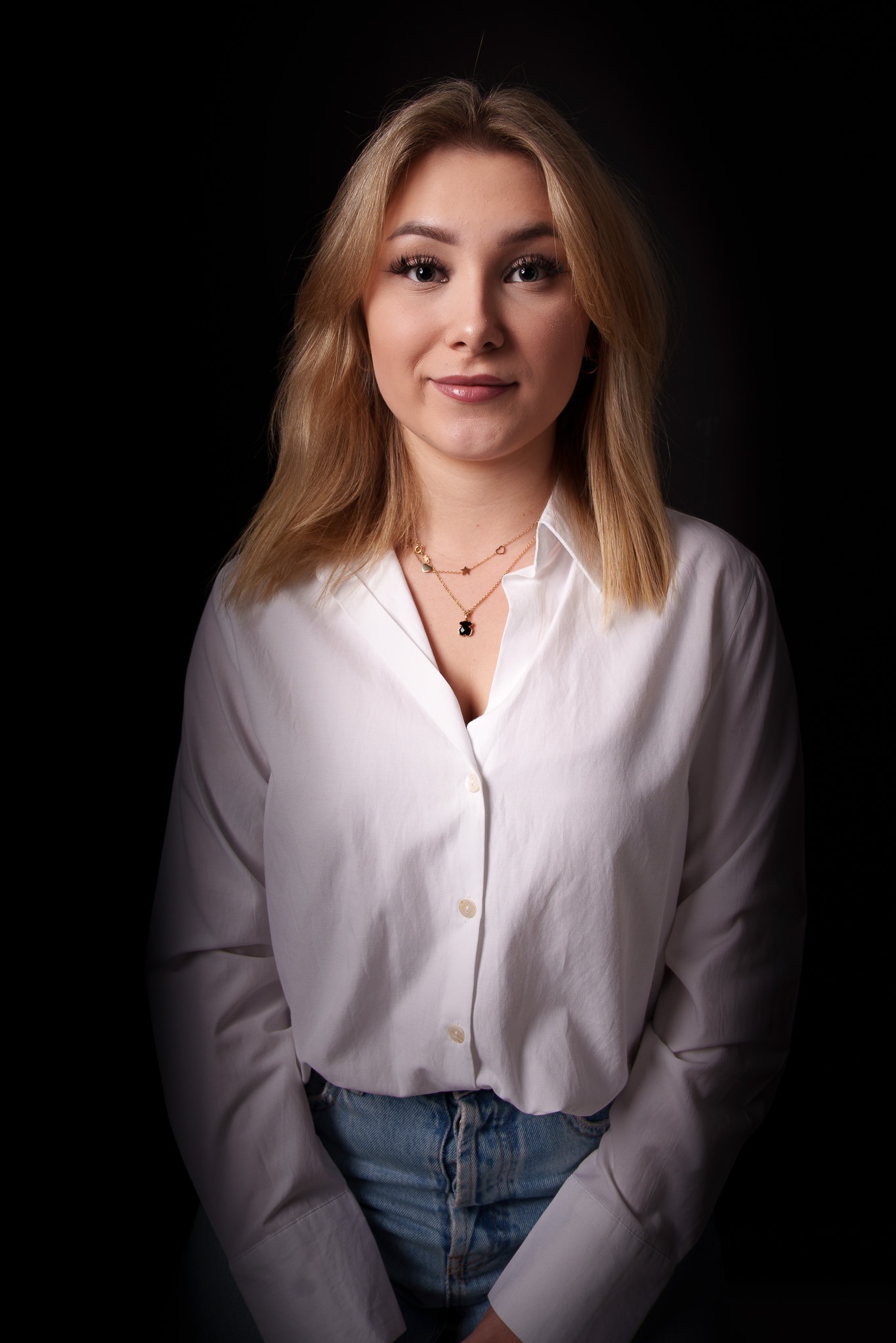 Portret młodej kobiety z blond włosami, ubranej w białą koszulę i jeansy, pozującej na ciemnym tle, z delikatnym uśmiechem i złotymi naszyjnikami.