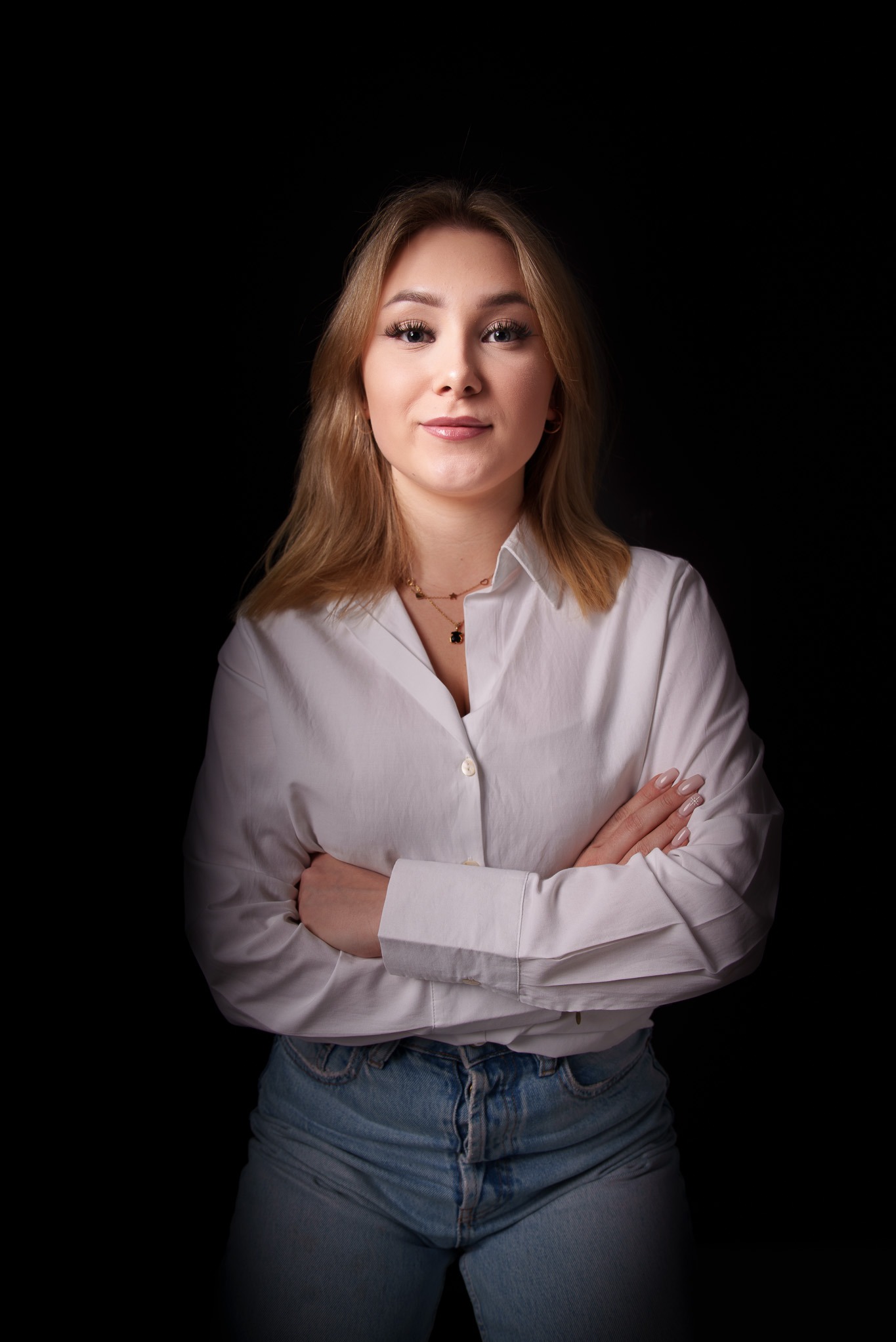 Portret kobiety w białej koszuli i jeansach na czarnym tle, z rękami skrzyżowanymi na piersiach, delikatny uśmiech, subtelny makijaż i złota biżuteria.