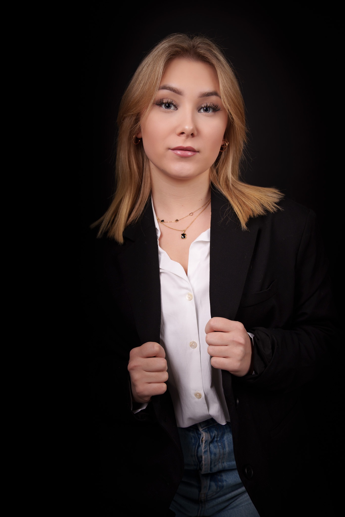 Portret młodej kobiety w czarnej marynarce i białej koszuli na ciemnym tle, z delikatnym makijażem i złotymi łańcuszkami na szyi.