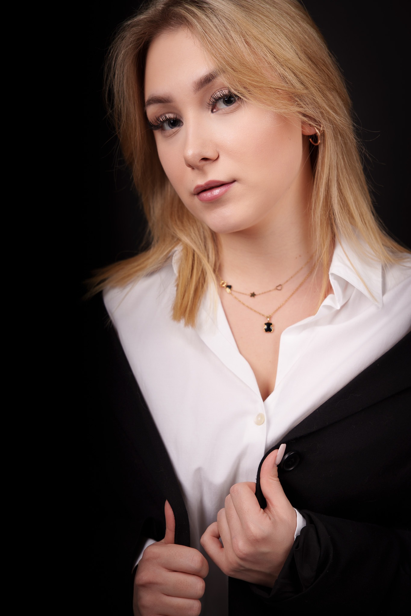 Portret kobiety z blond włosami w białej koszuli i czarnej marynarce na czarnym tle, z delikatnym makijażem i złotymi łańcuszkami z zawieszkami.