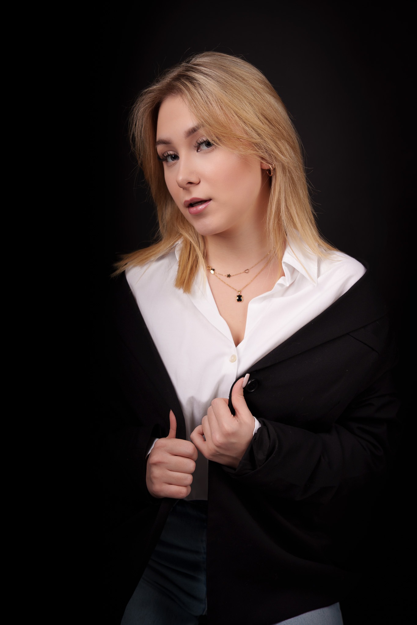 Portret młodej kobiety z blond włosami w białej koszuli i czarnej marynarce na czarnym tle, z delikatną biżuterią na szyi.