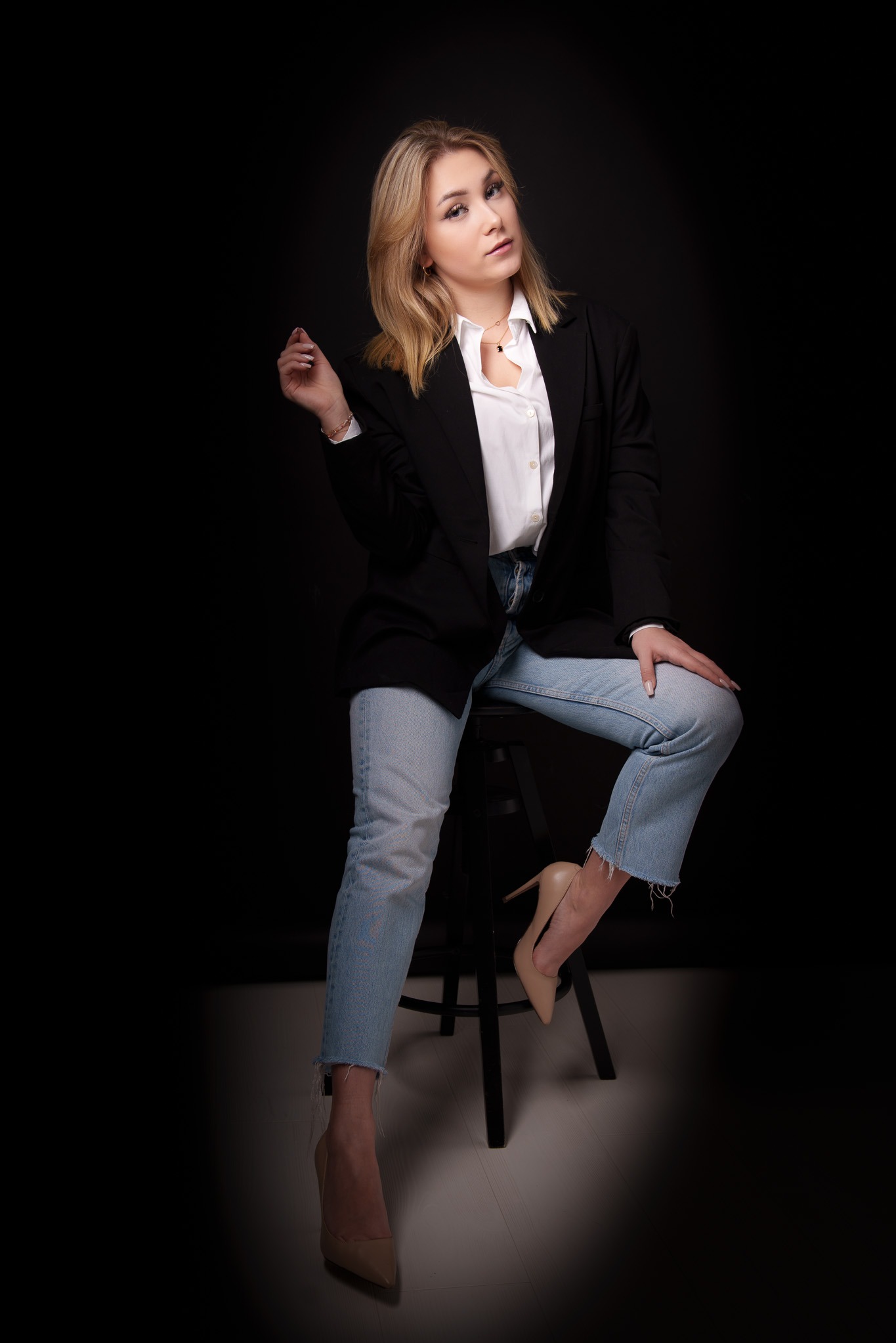 Portret młodej kobiety siedzącej na stołku, ubrana w czarną marynarkę, białą koszulę i jasne jeansy, pozującej na ciemnym tle.